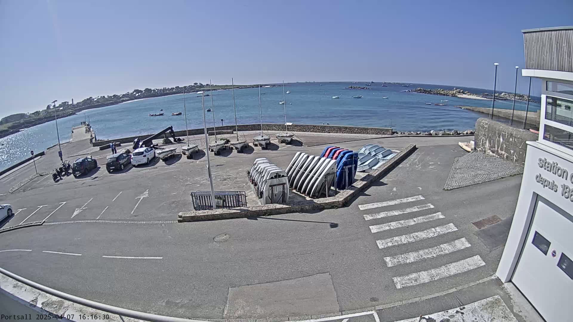 A sunny day at a harbor shows parked cars near a building and numerous small sailboats stored on land next to calm blue water.
