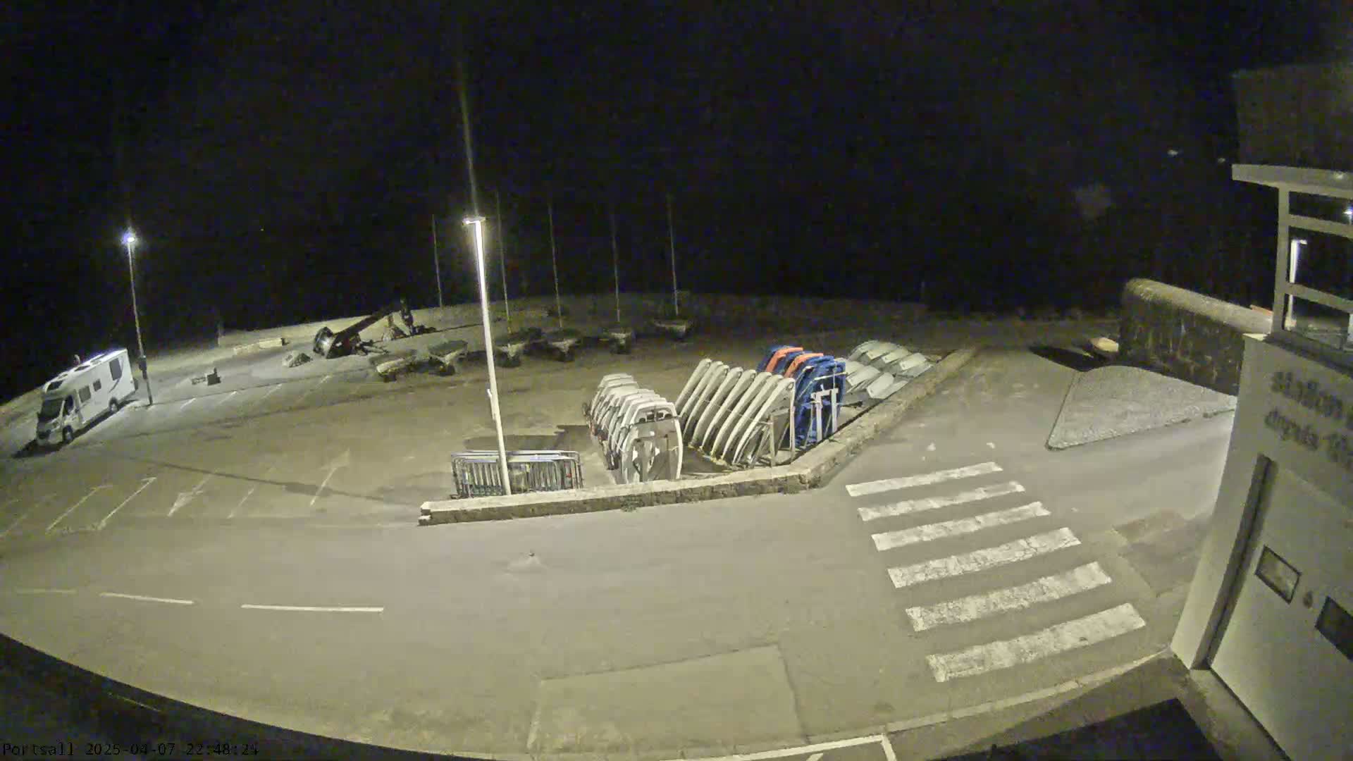 A nighttime, outdoor view of an empty parking lot with several boats stored upright on their sides, a campervan, and a crosswalk.