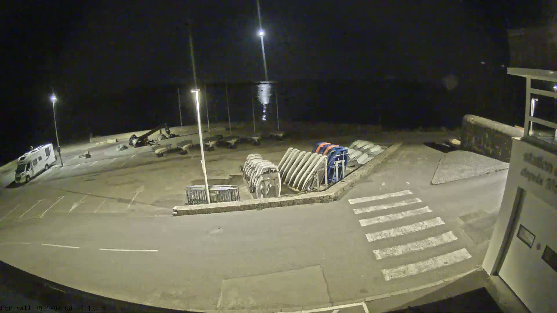 A nighttime view of an empty parking lot near a body of water, with several boats or boat hulls stored nearby, under a mostly clear sky with a bright moon.
