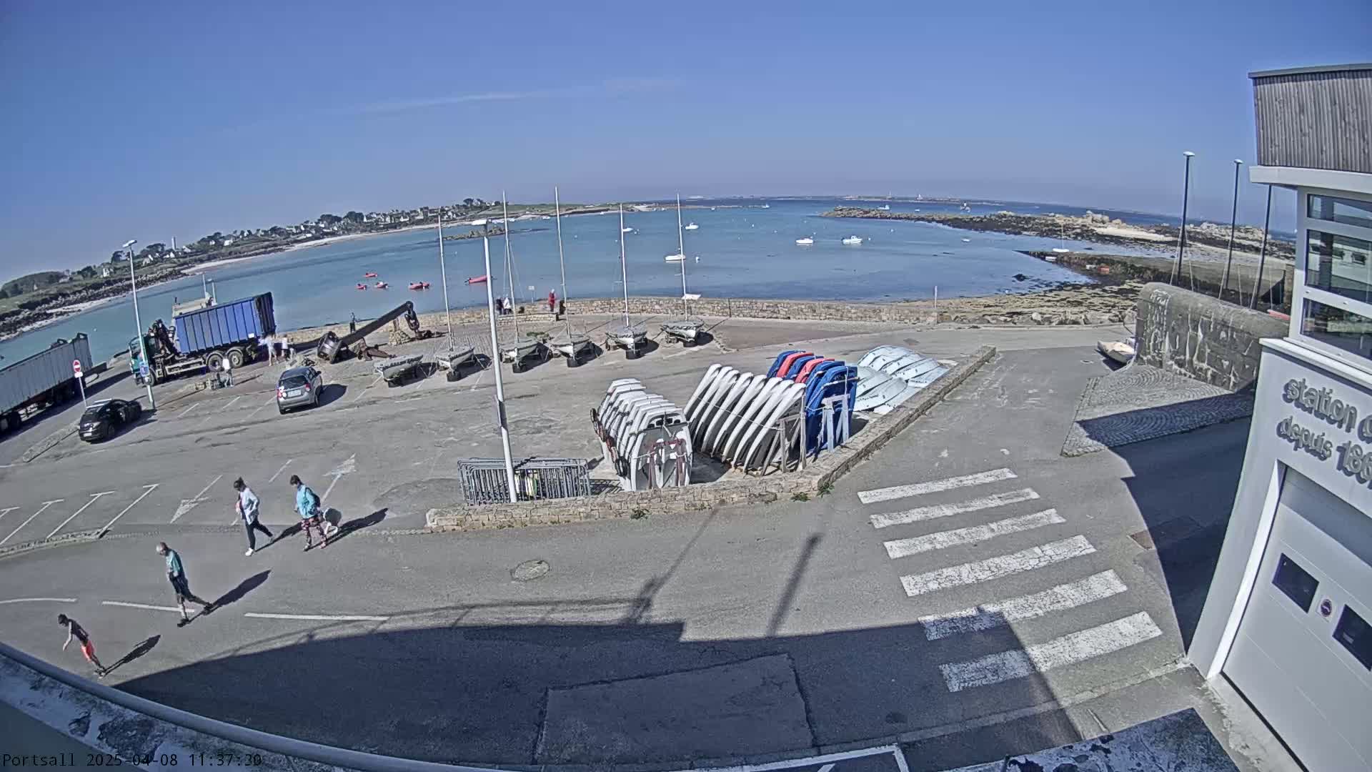 A sunny day at a harbor shows several parked cars, a few people walking, numerous sailboats stored upright, and a couple of trucks near a body of calm water.