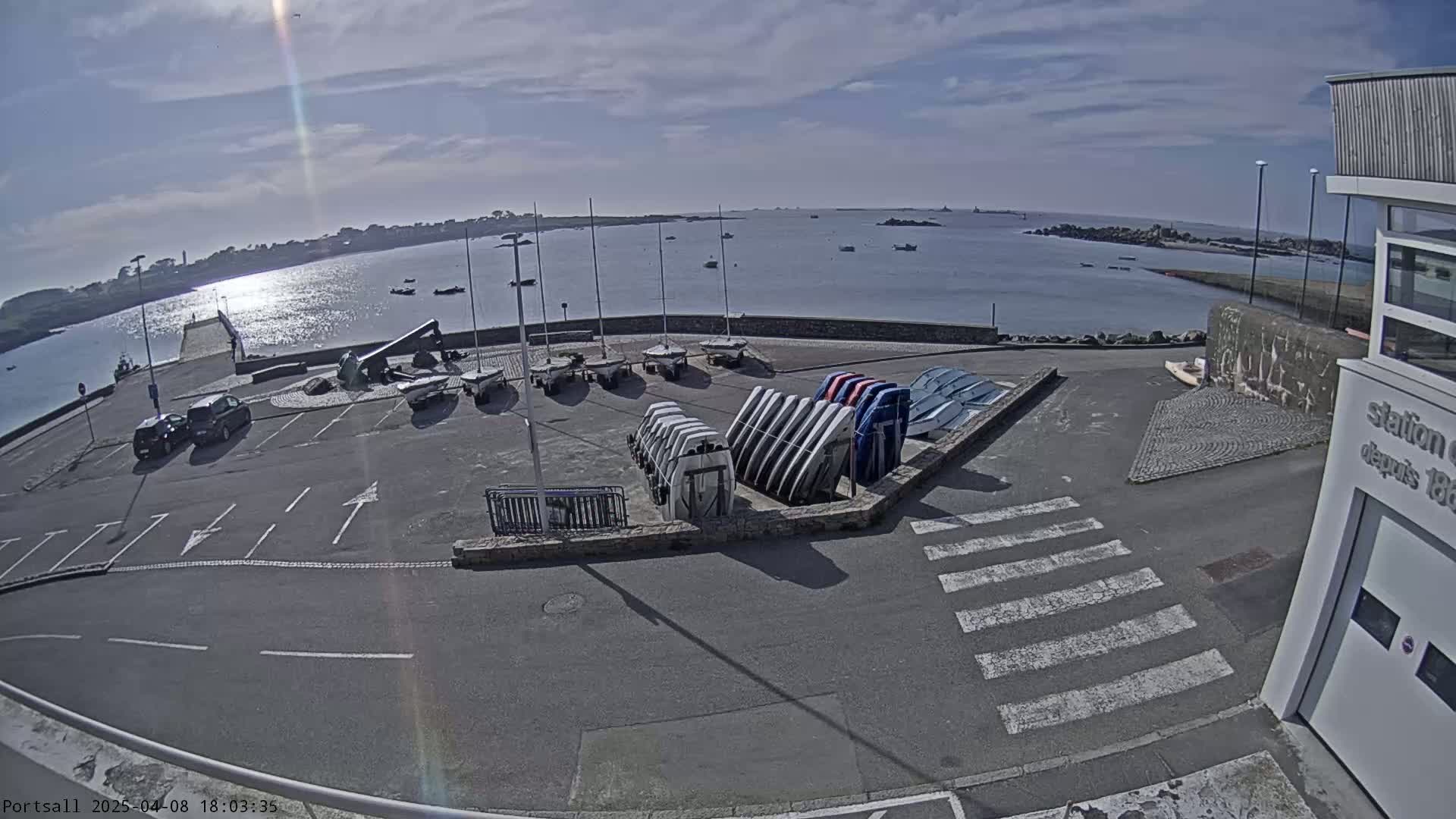 A sunny day at a harbor shows a parking lot with several small boats and cars, overlooking a calm body of water with more boats visible in the distance.