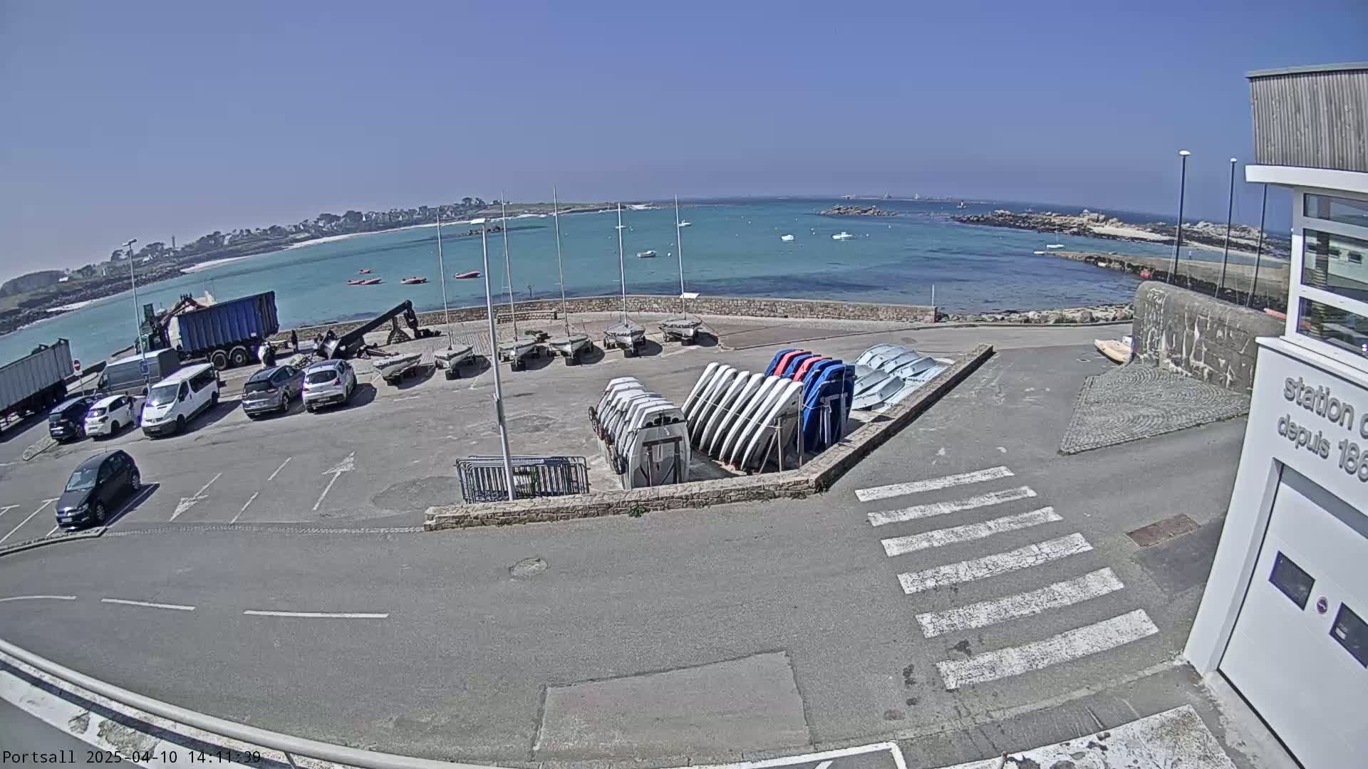 A sunny day at a harbor shows several cars parked near a building, with numerous sailboats stored on racks and others in the water.
