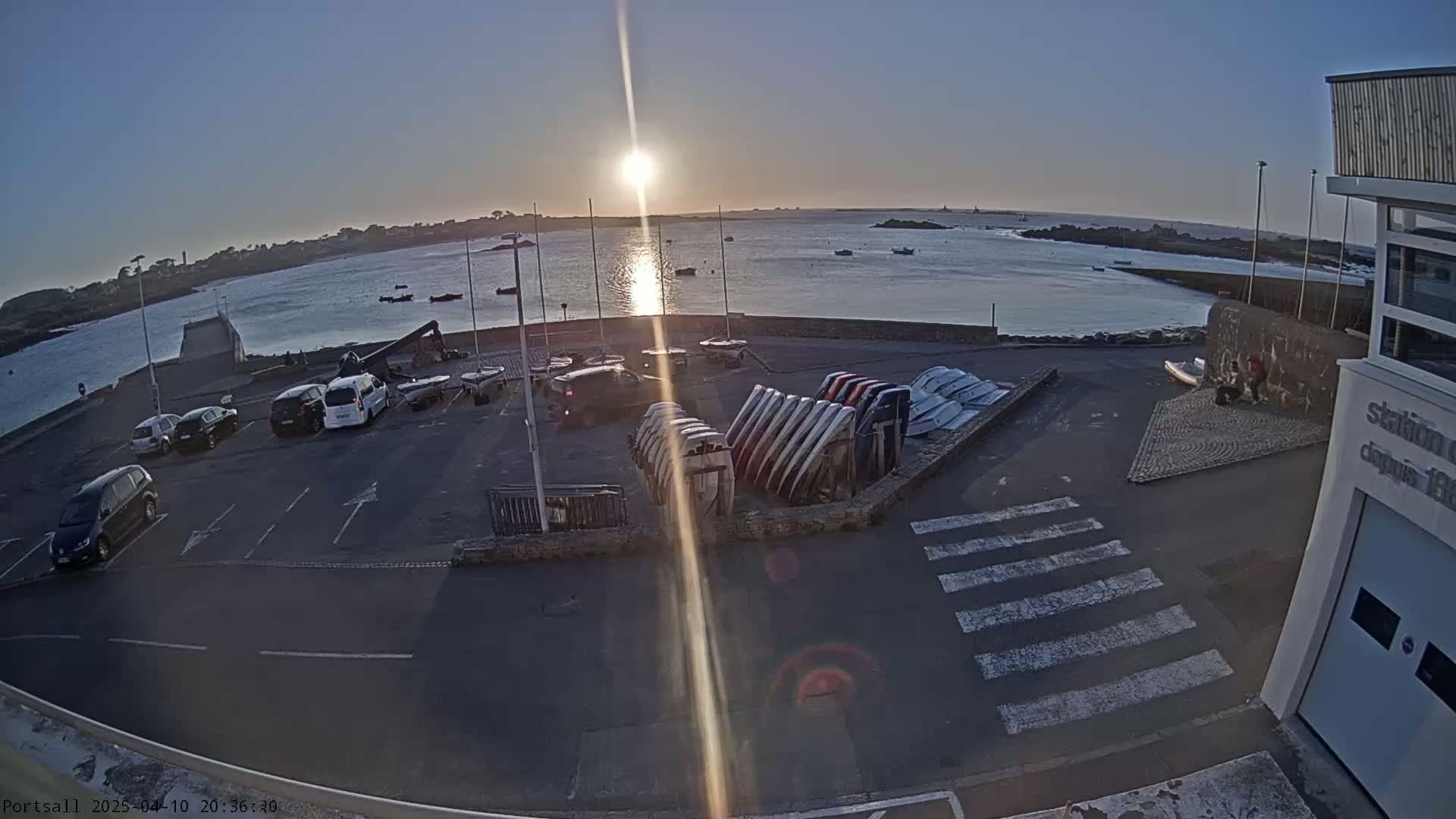 A parking lot with several cars and numerous small boats stacked together overlooks a calm body of water at sunset.