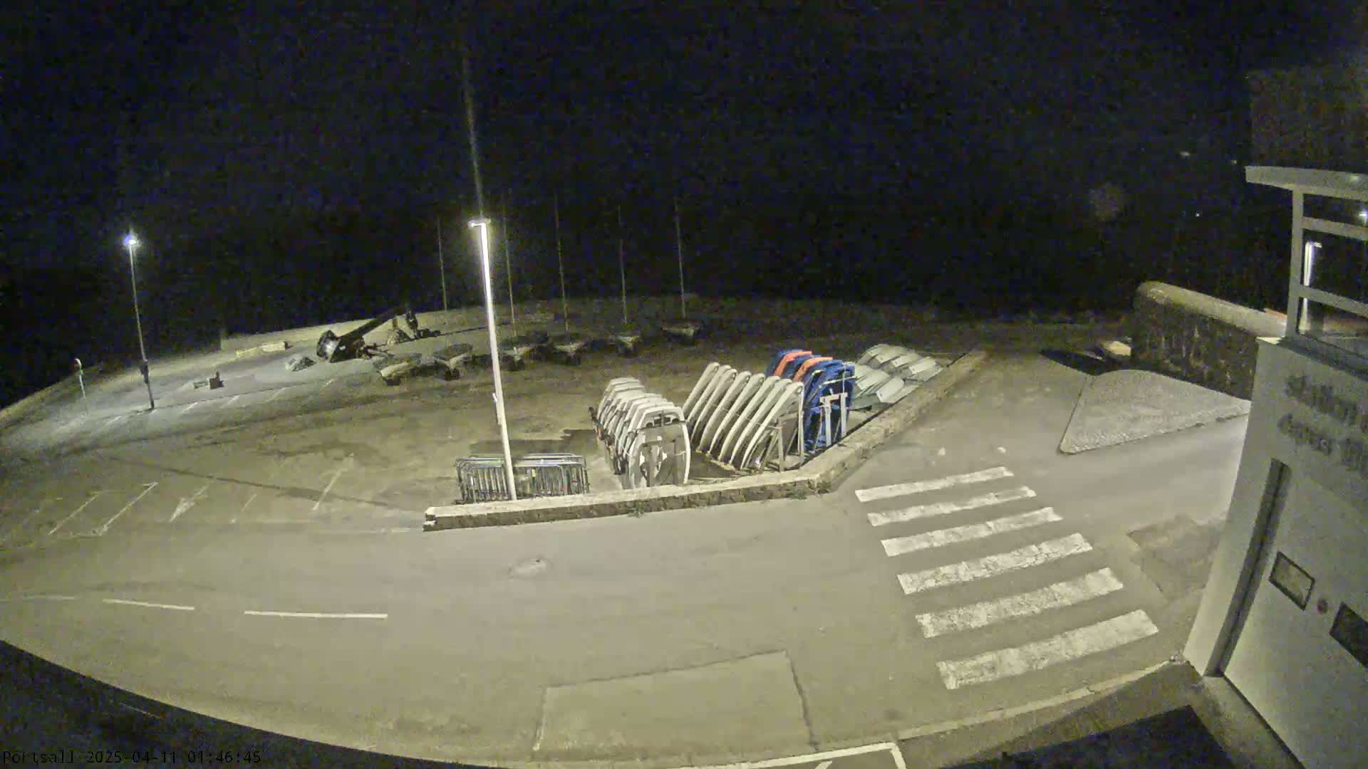 A nighttime, outdoor view of an empty parking lot with several boats stored on their sides, near a crosswalk and building.