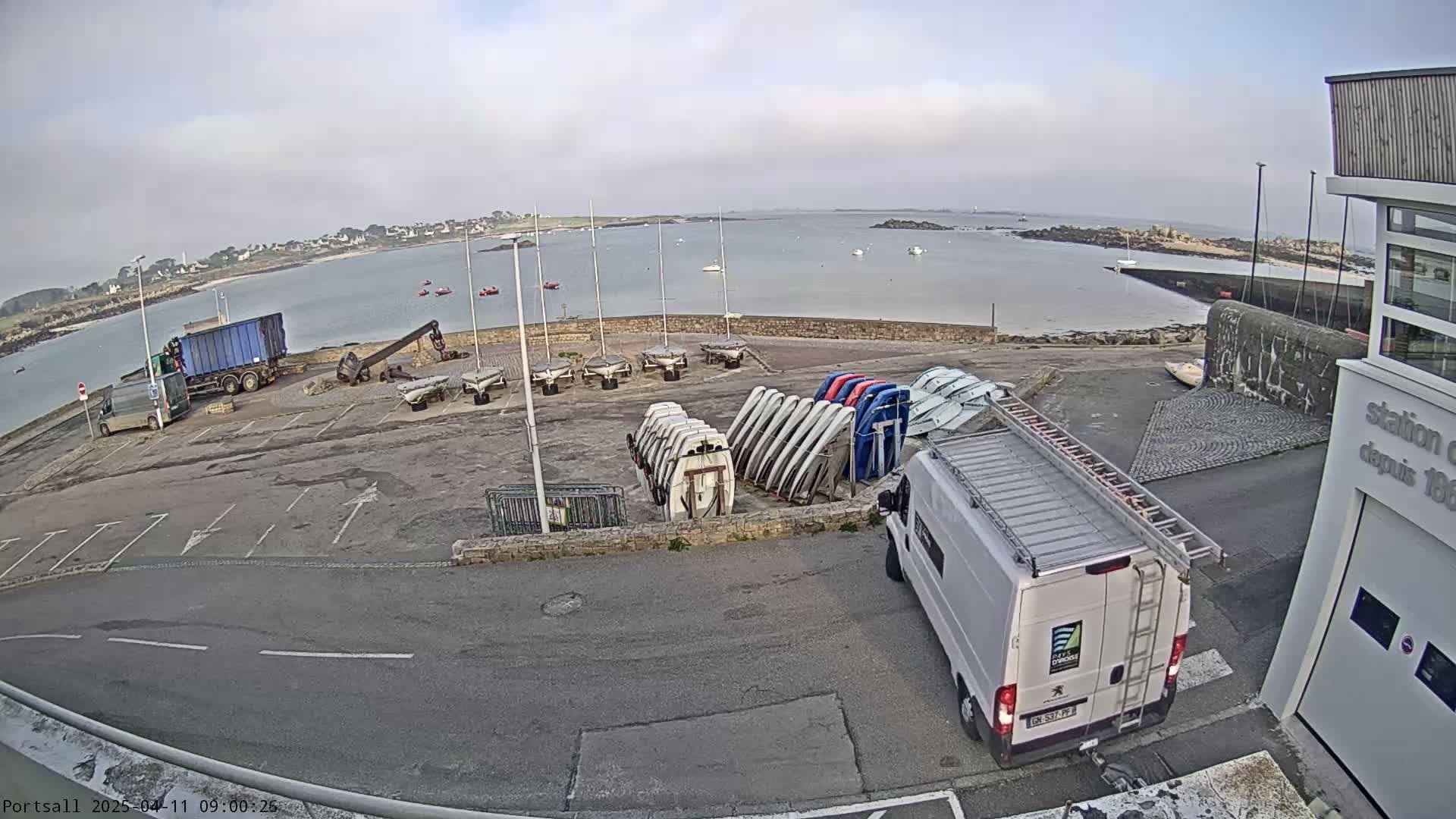 A harbor scene on an overcast day shows a parking lot with several boats, a van, and a truck, all near a building and a calm body of water with a few small boats visible in the distance.