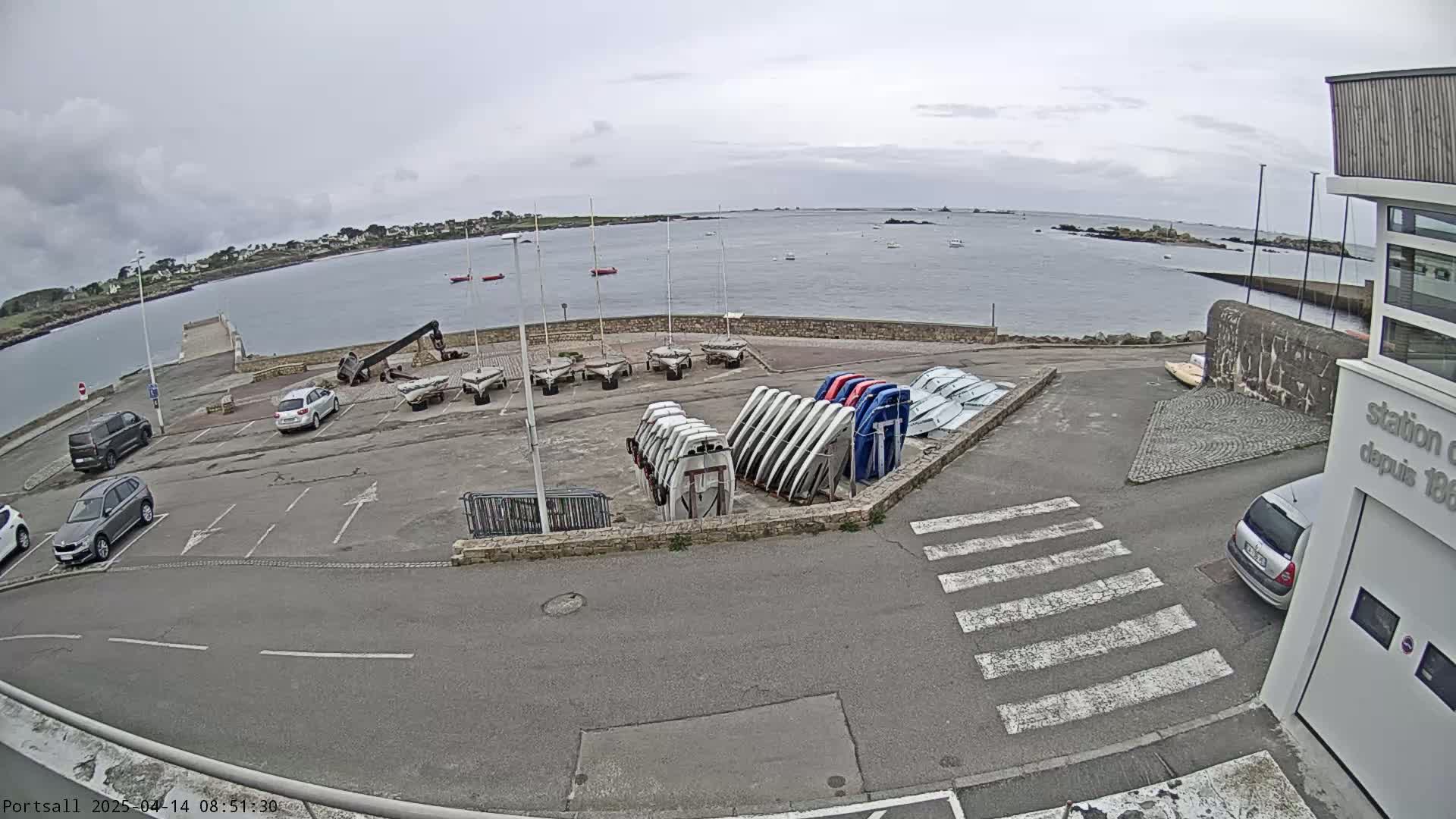 A parking lot with several cars and numerous small boats stored upright on racks overlooks a calm body of water under an overcast sky.