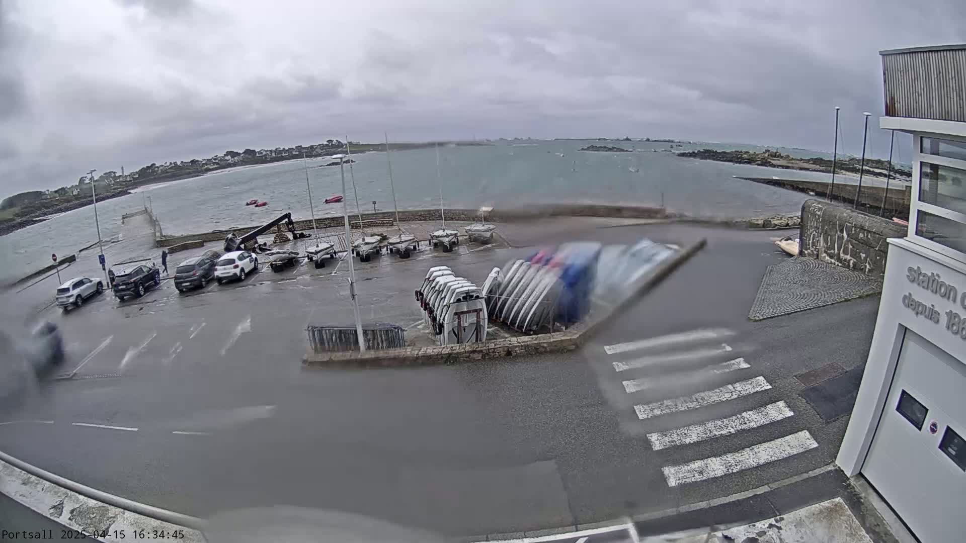 A parking lot with several cars and numerous small sailboats stacked on trailers is next to a body of water under an overcast sky, with rain visible on the camera lens.