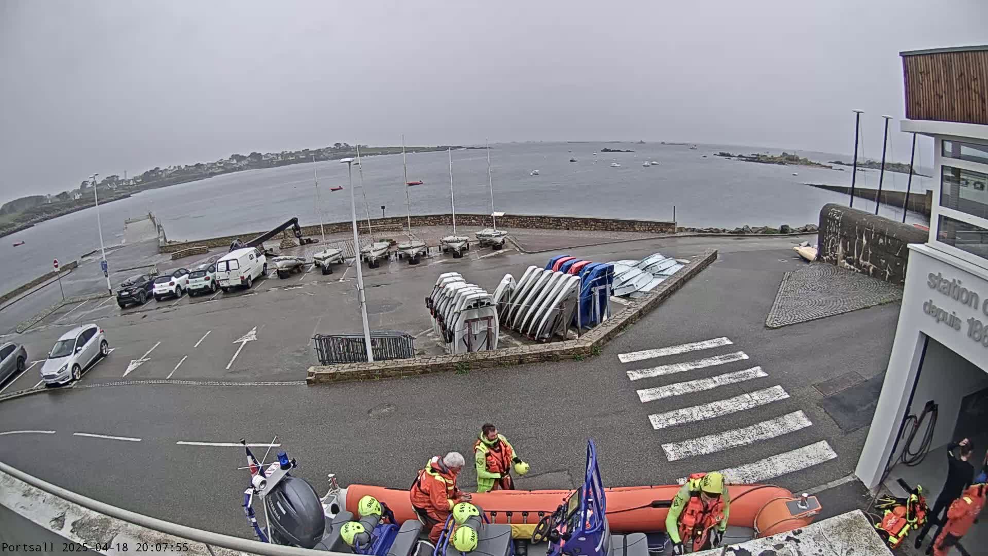 A parking lot with several cars and sailboats, next to a body of water under an overcast sky, with rescue workers preparing a rescue boat.