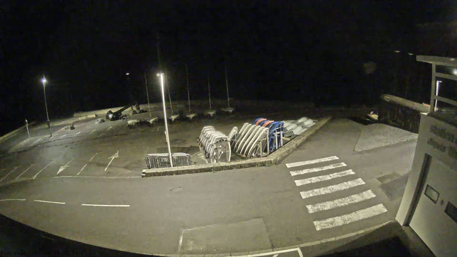 An empty parking lot at night, next to a building and a row of upturned boats under lights.