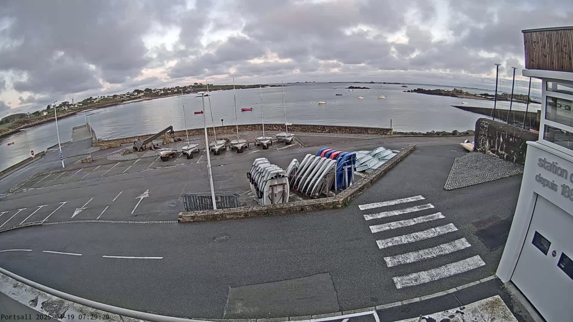 A parking lot with numerous sailboats stored upright, near a calm body of water under an overcast sky.
