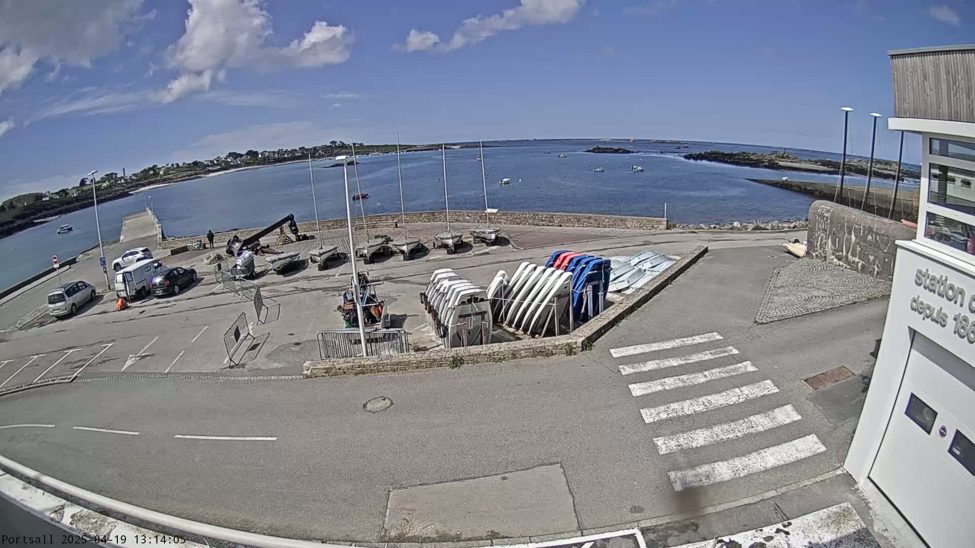 A sunny day at a harbor shows several parked cars, a small crane, numerous sailboats and other small watercraft, and a building near a calm body of water.