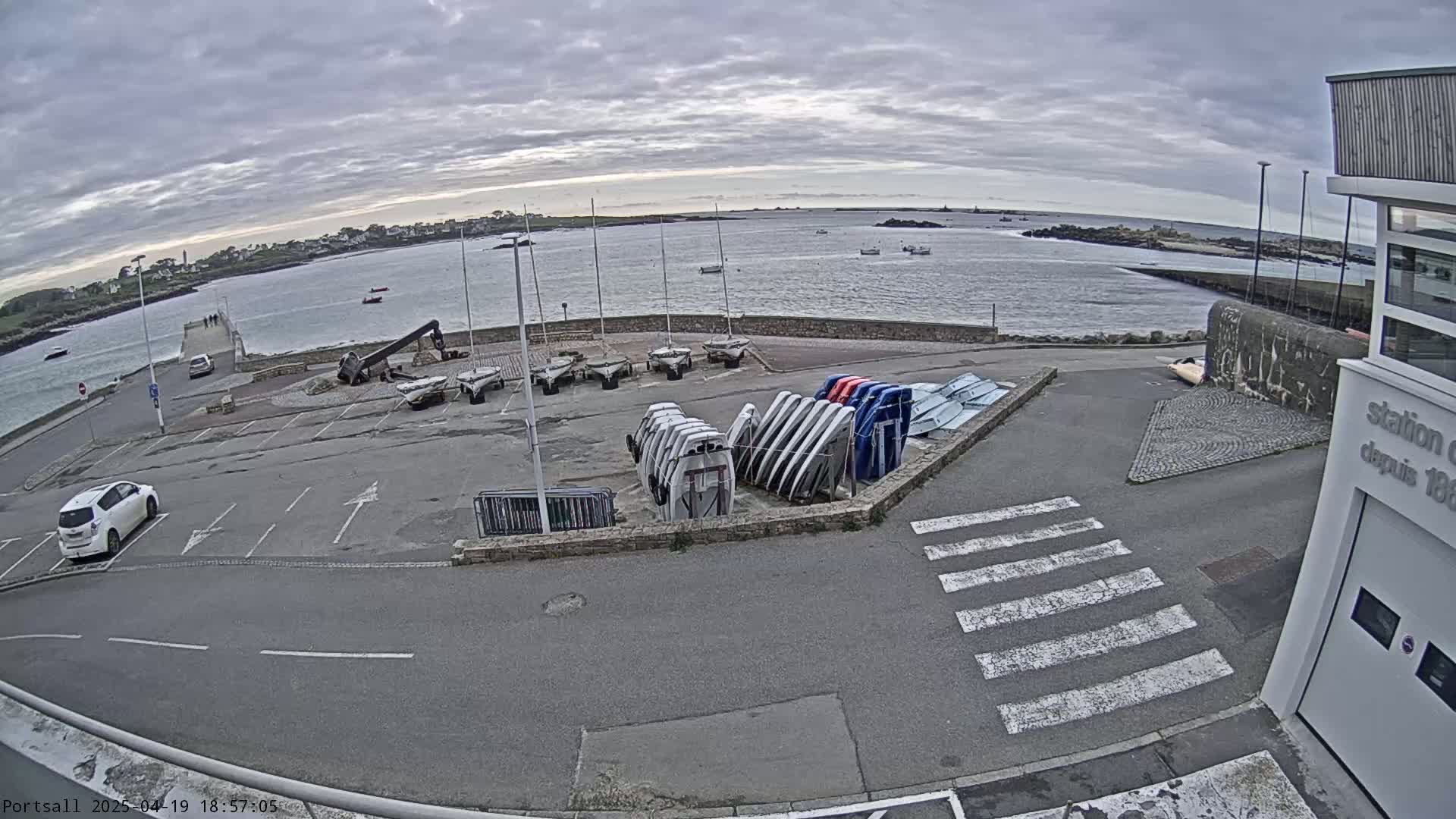 A harbor scene on an overcast day shows sailboats stored on land, a few boats on the water, and a car parked in an empty lot next to a building.