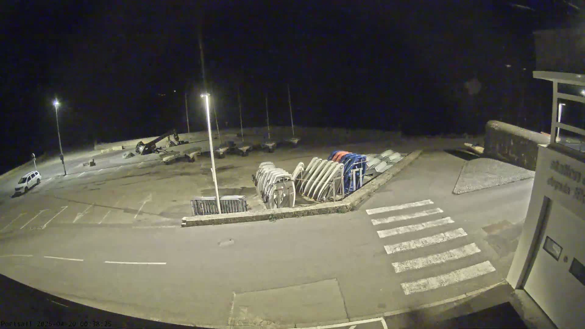 A nighttime, outdoor view of an empty parking lot with several boats stored upright near a building and a crosswalk.