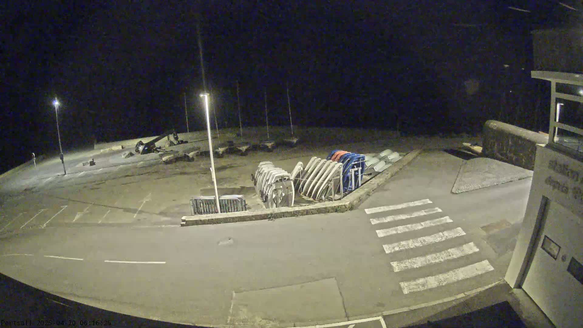 A nighttime, outdoor view shows an empty parking lot with several boat hulls stored upright against a low wall, under illuminated streetlights.