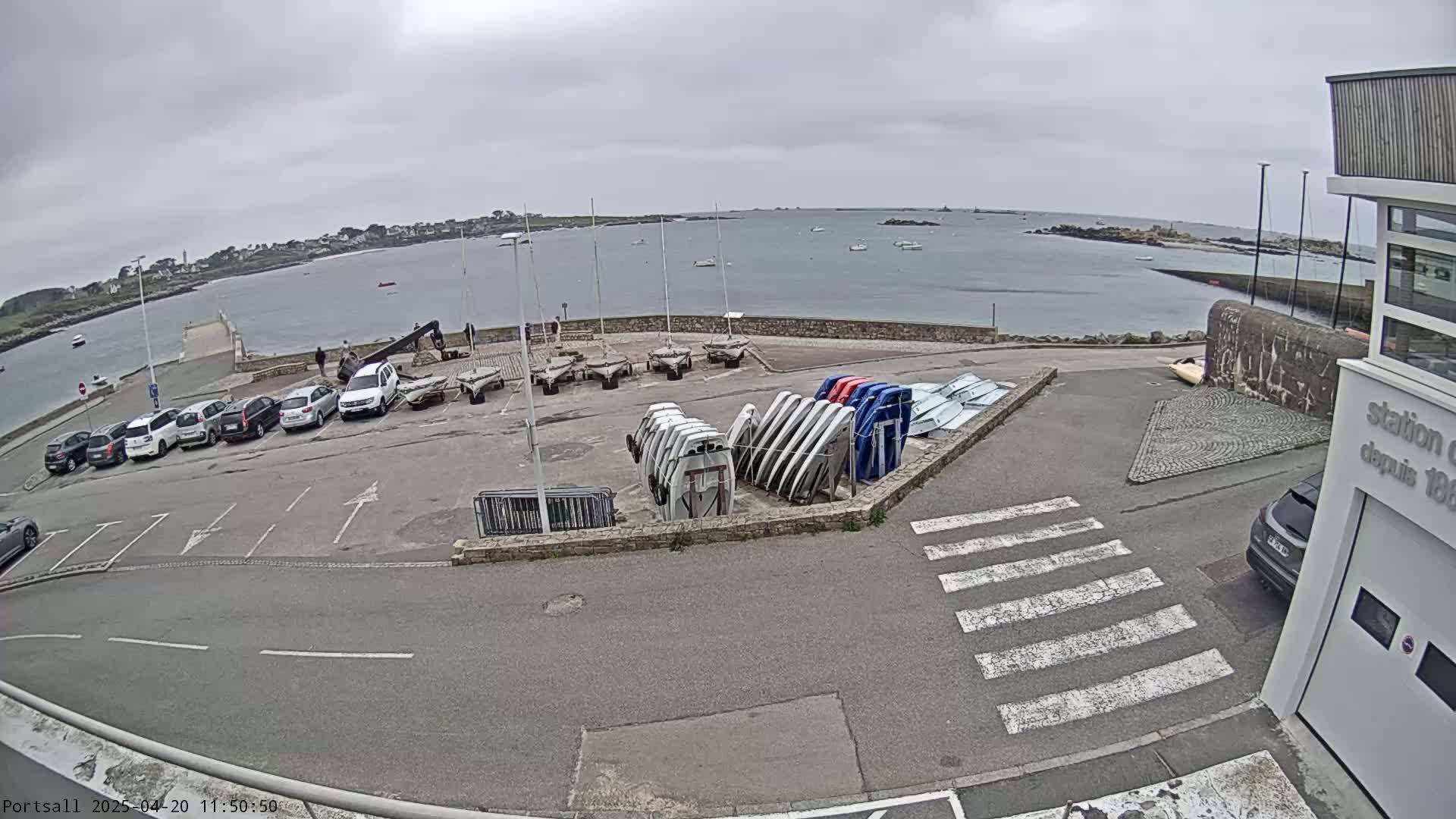 A parking lot with several cars and numerous small boats stacked together overlooks a calm body of water under an overcast sky.