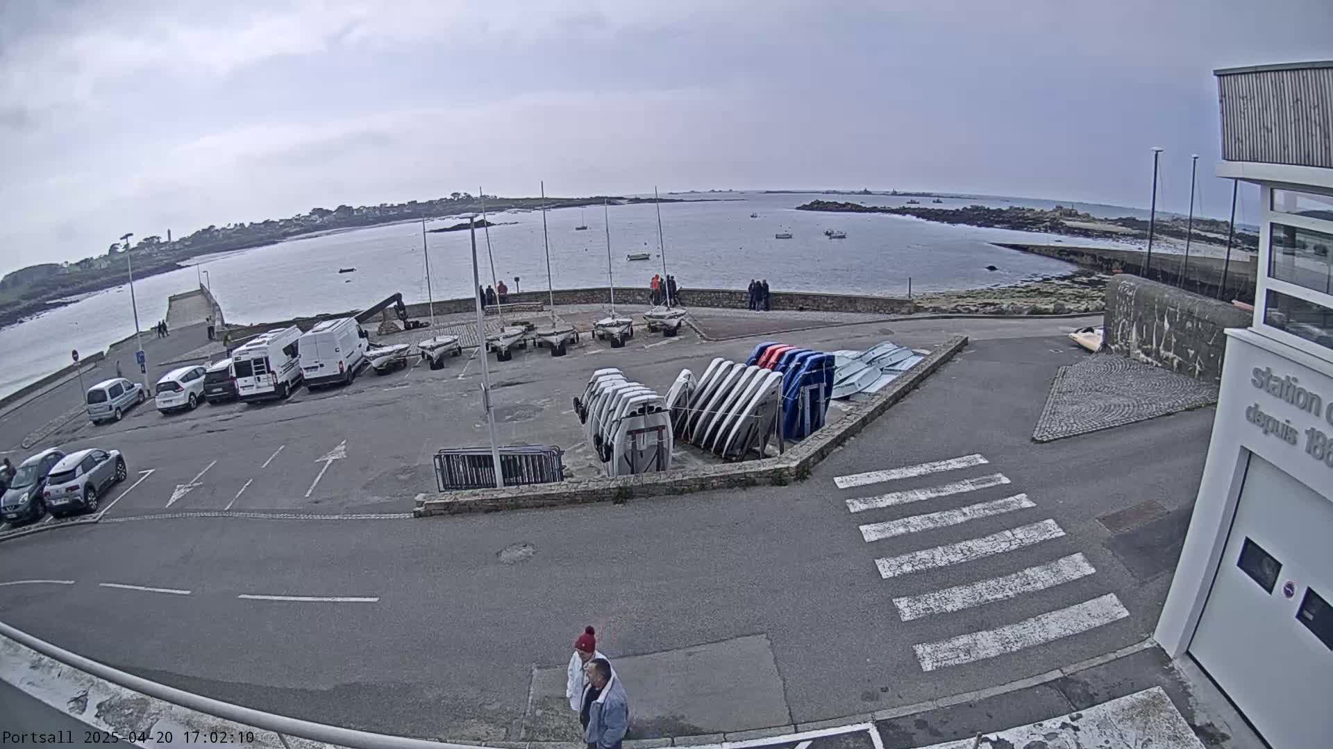 A parking lot with several cars and numerous small boats stored upright is situated near a calm body of water under an overcast sky.