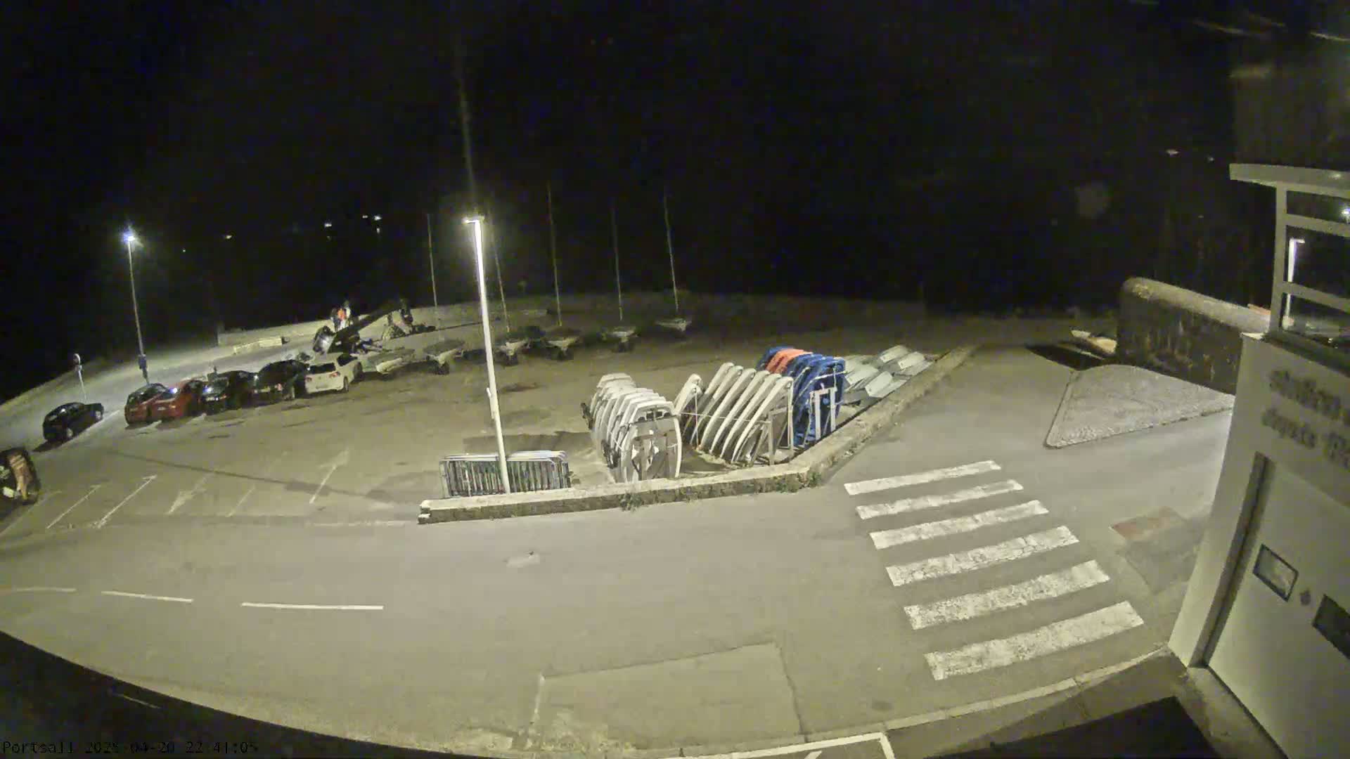 A nighttime, outdoor view of a parking lot with several cars parked, next to a boat storage area with numerous upturned boats, all under artificial lighting.