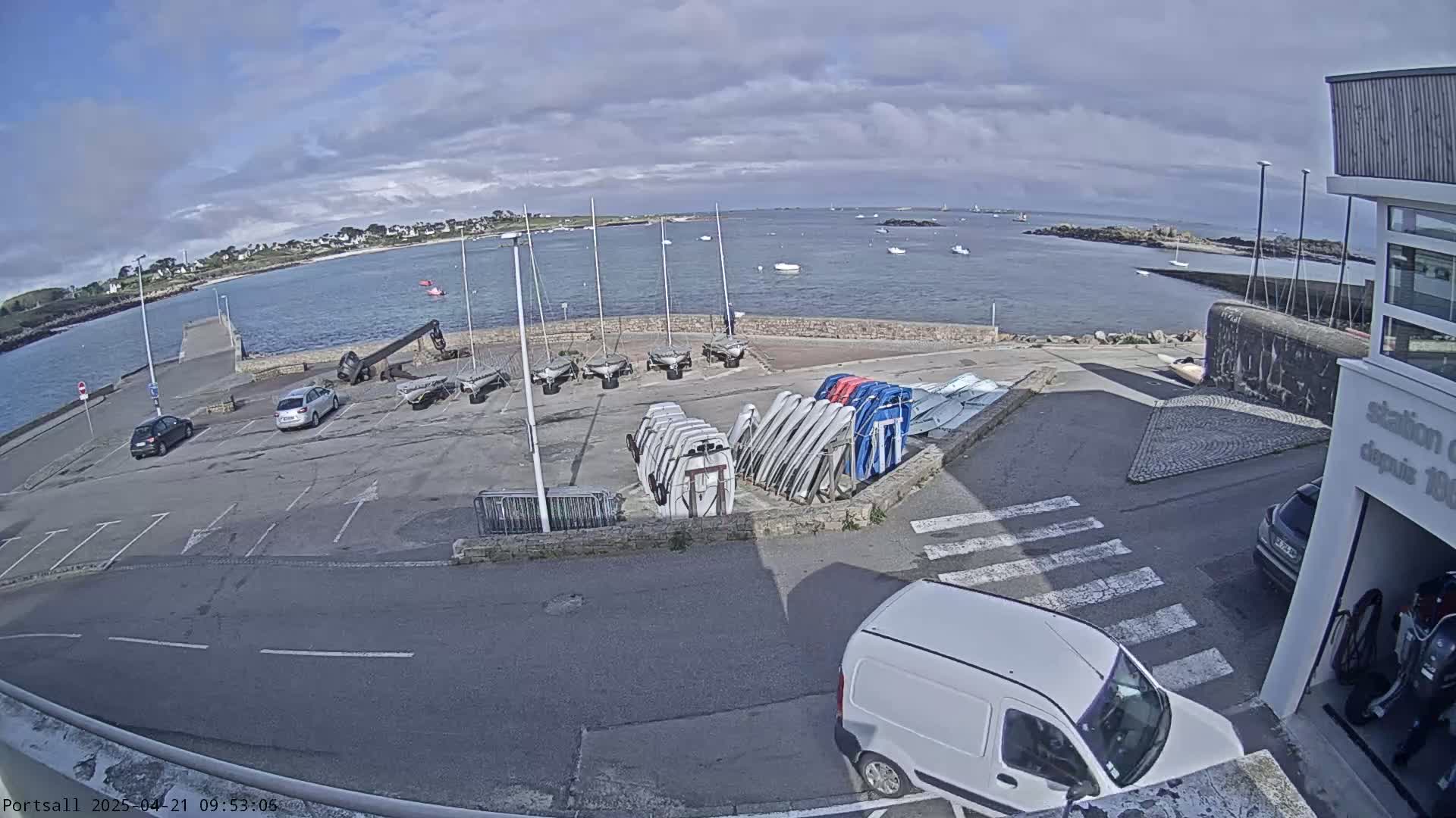 A partly cloudy day at a harbor with several sailboats and cars parked in a lot near a building, with a white van in the foreground.