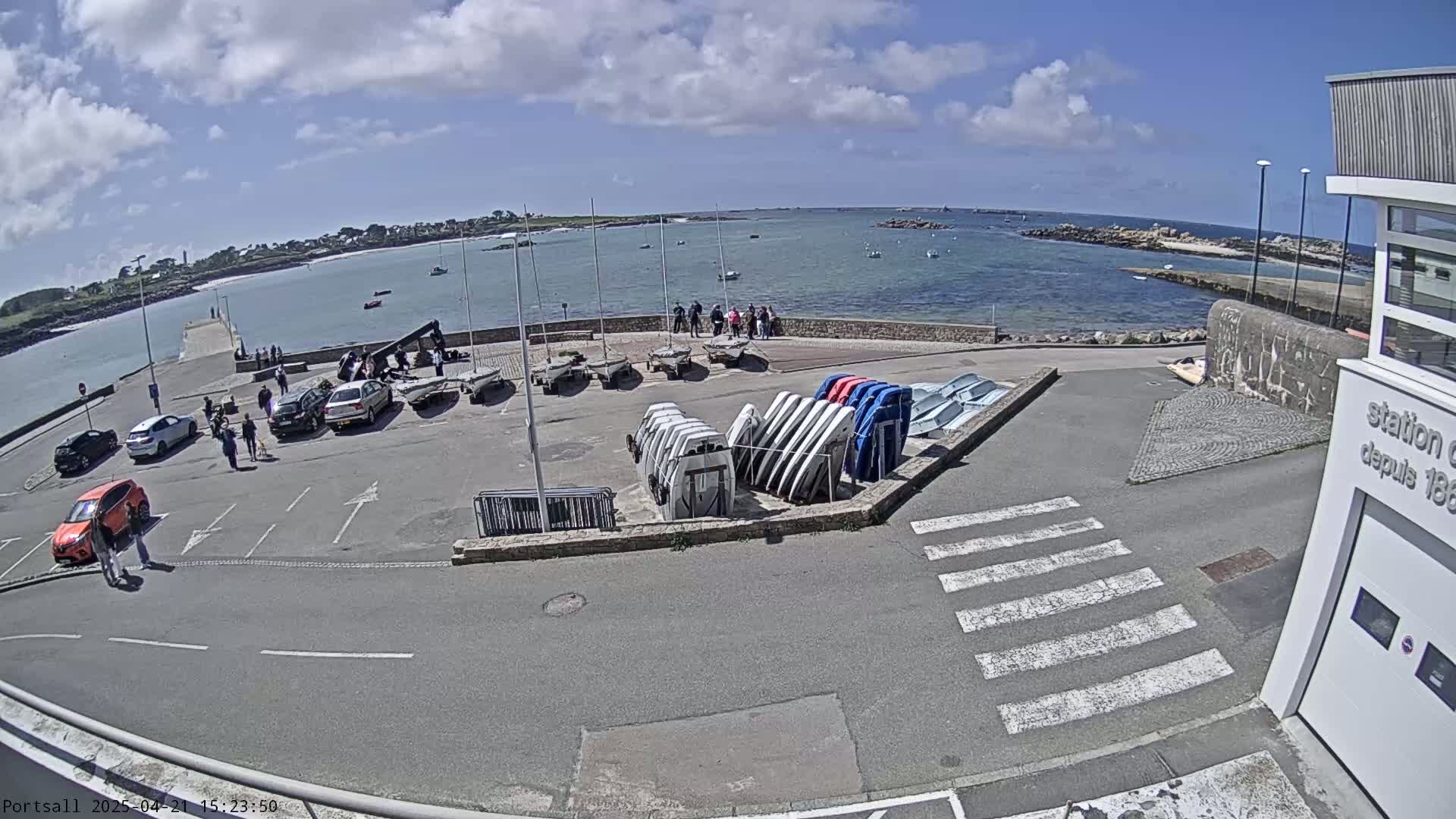 A sunny day at a harbor shows several cars parked near sailboats and small motorboats, with people walking around near the water.