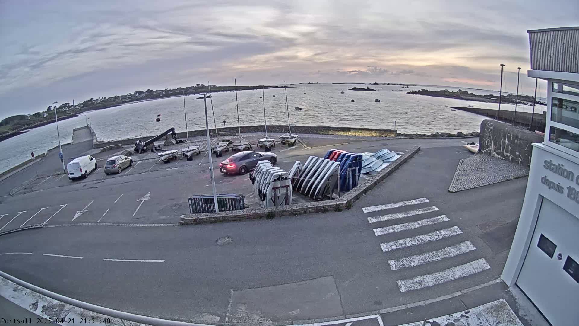 A parking lot with several cars and numerous small boats stored on racks overlooks a calm body of water under a cloudy sunset sky.