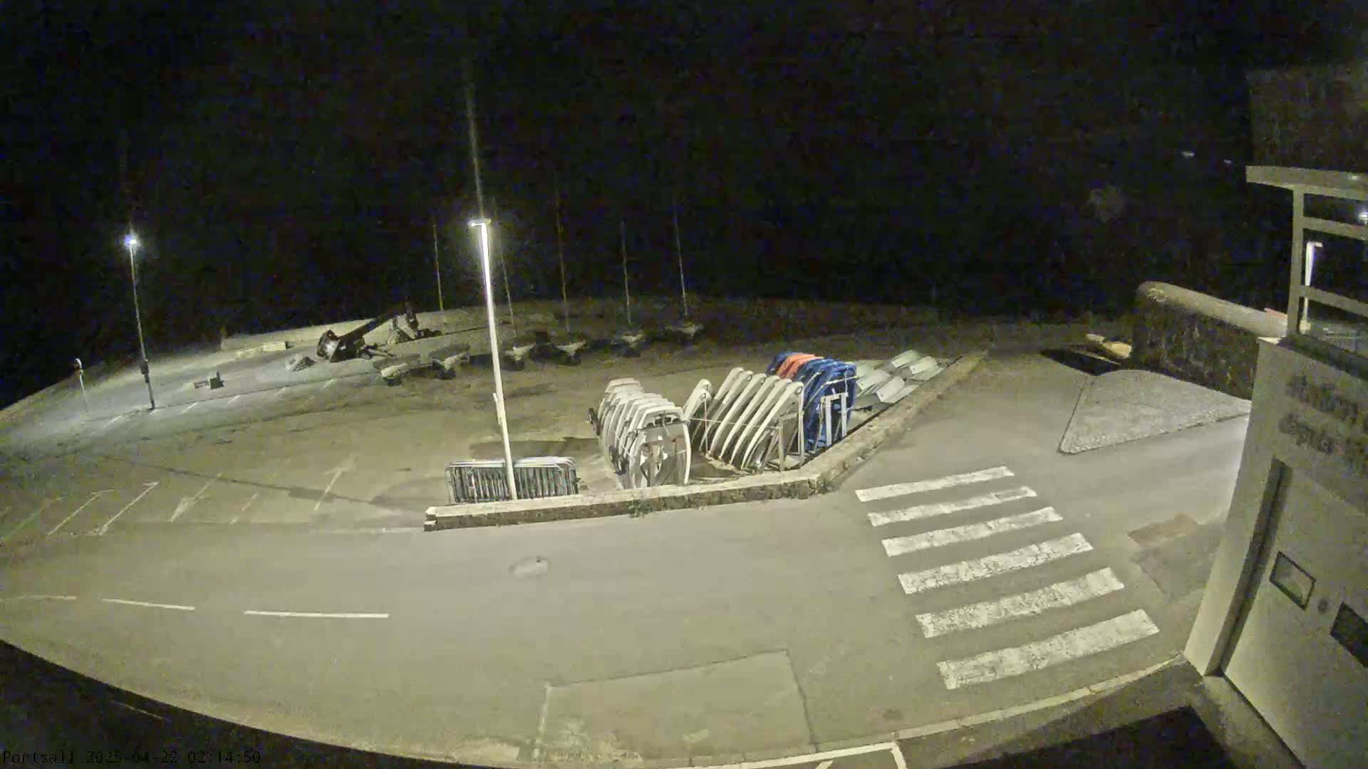At night, a mostly empty parking lot contains several boats stored upright, next to a crosswalk.