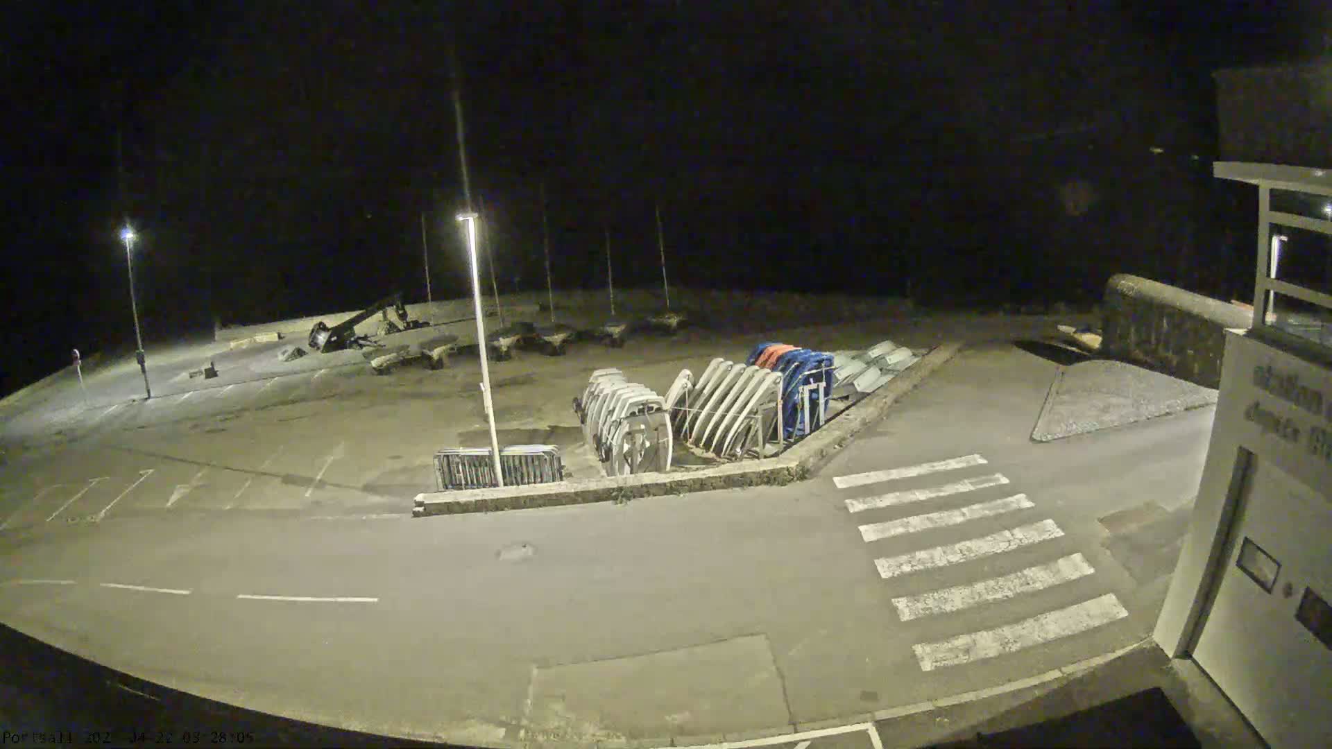 A nighttime, outdoor view of an empty parking lot with several boat hulls stored upright along a wall, and a crosswalk leading to a building.
