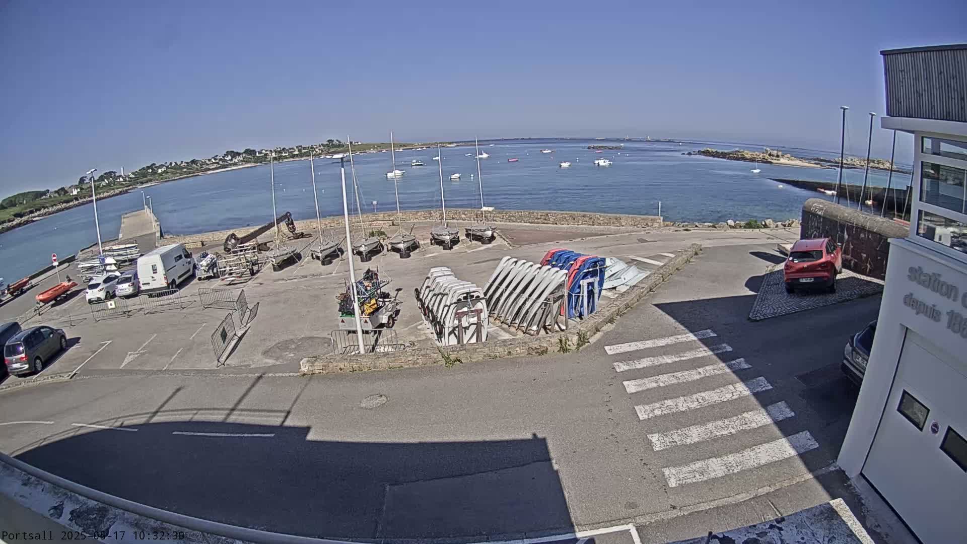 A sunny day at a harbor shows numerous small sailboats docked and stored, along with several cars parked in a lot near a building.
