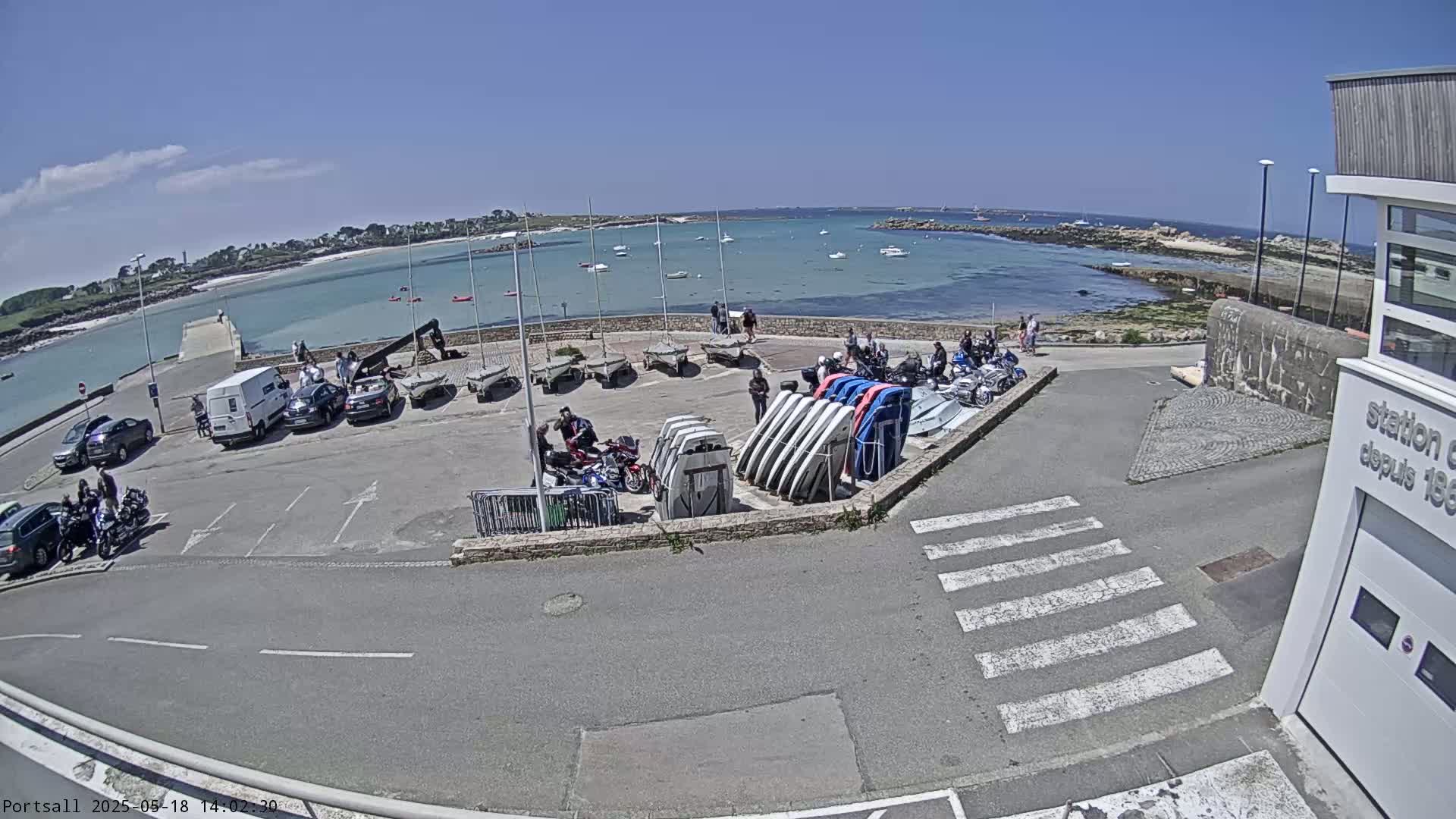 A sunny day at a harbor shows several parked cars and motorcycles near a collection of sailboats and small motorboats, with a calm body of water and distant islands visible in the background.