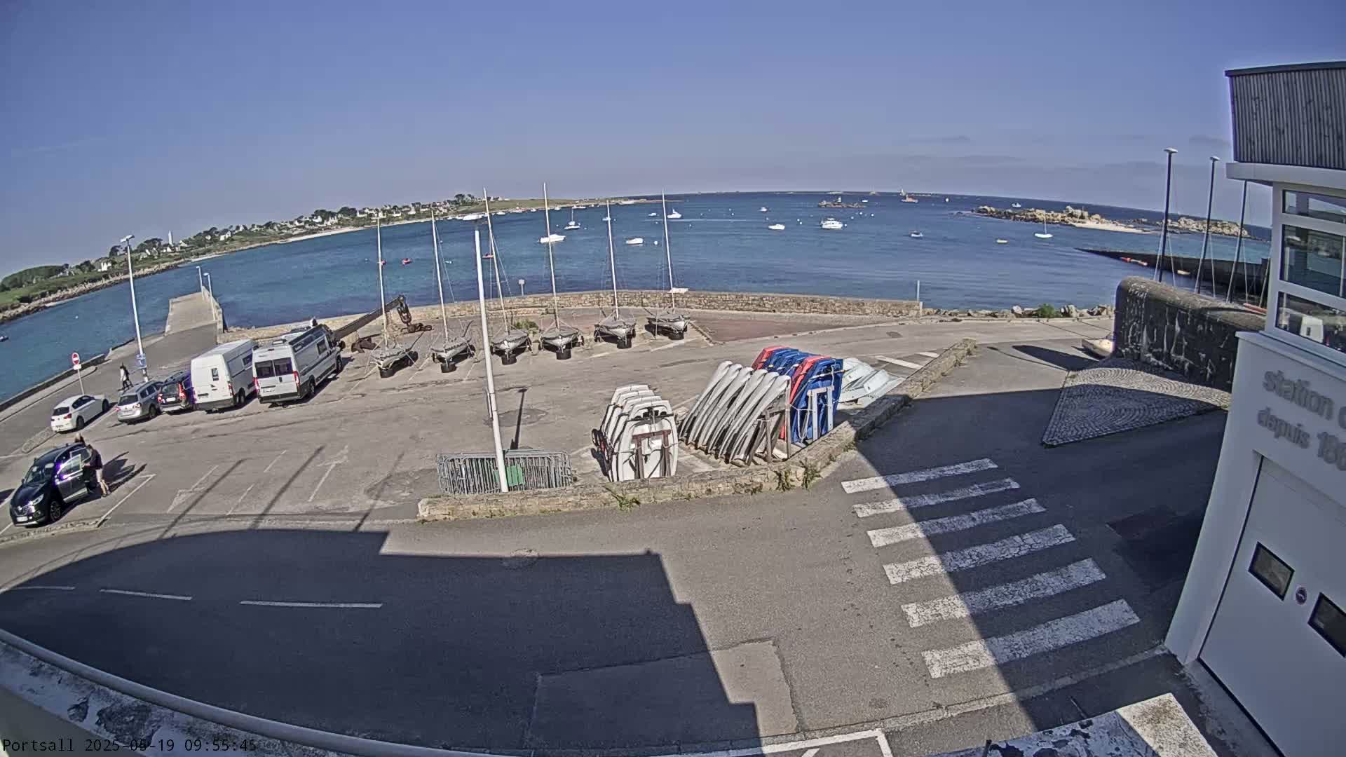 A sunny day overlooking a harbor with parked cars, several sailboats, and numerous small boats stored on racks.