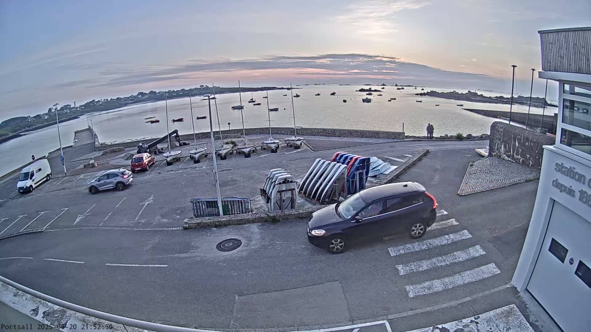A parking lot with several cars and numerous sailboats stored upright is situated next to a calm body of water at sunset.