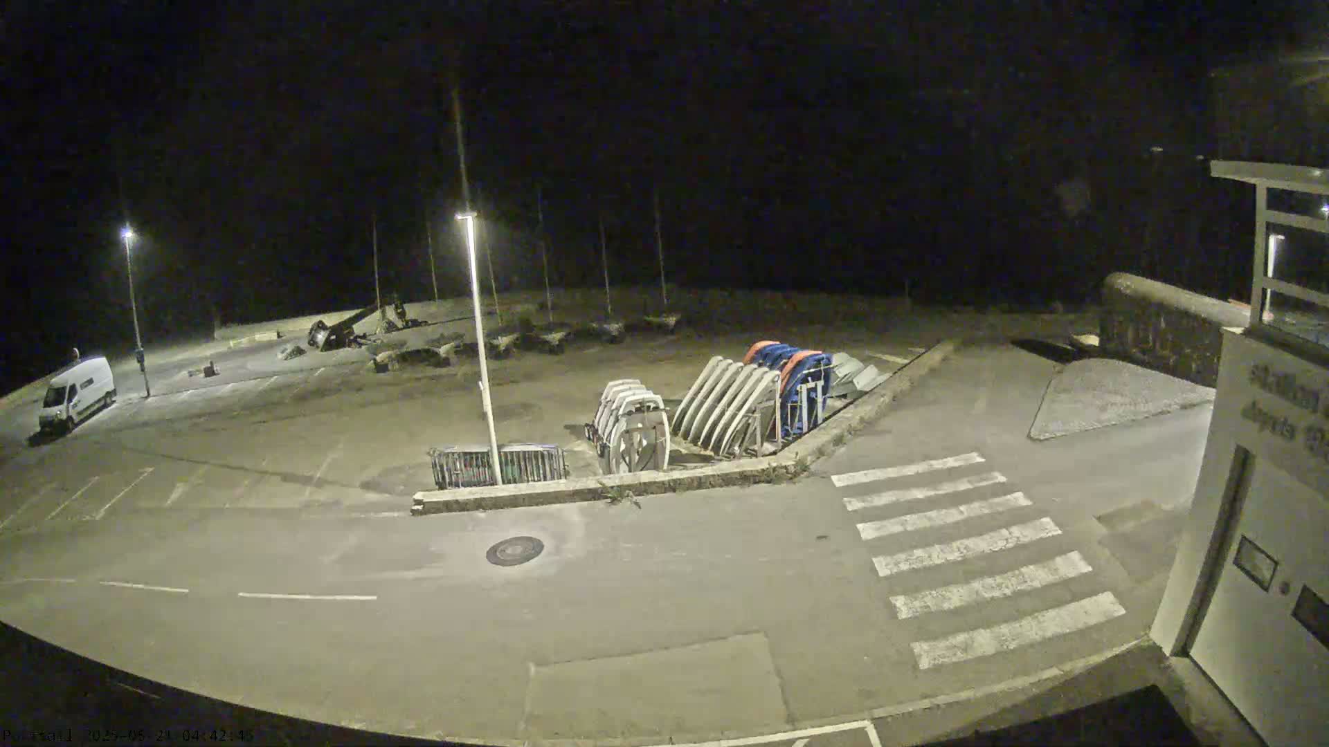 A nighttime view of an empty parking lot with several boats stored on stands, a white van, and a crosswalk.
