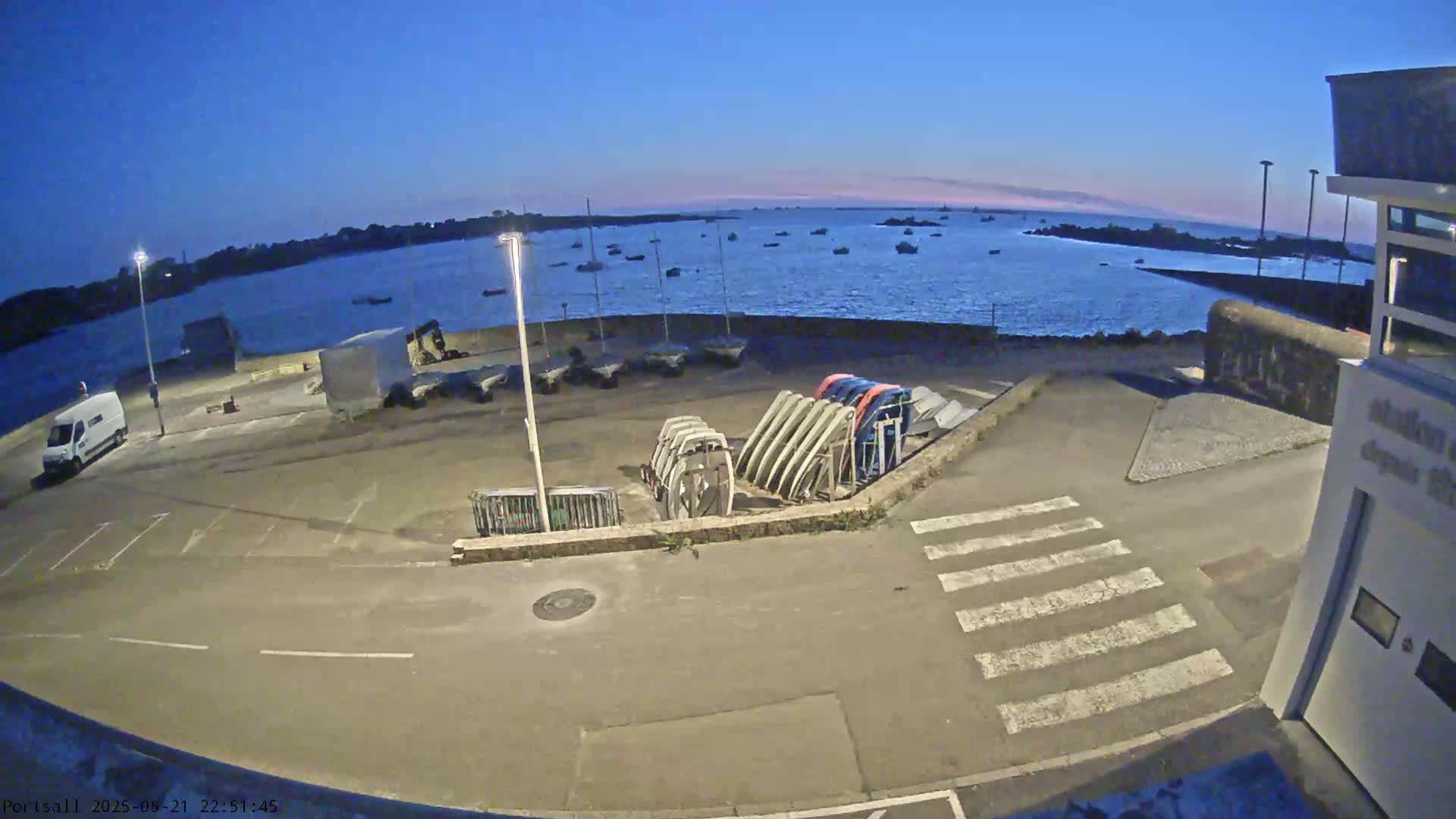 A calm, twilight-lit harbor scene shows a parking lot with several boats and boat trailers, next to a body of water with other boats visible in the distance.