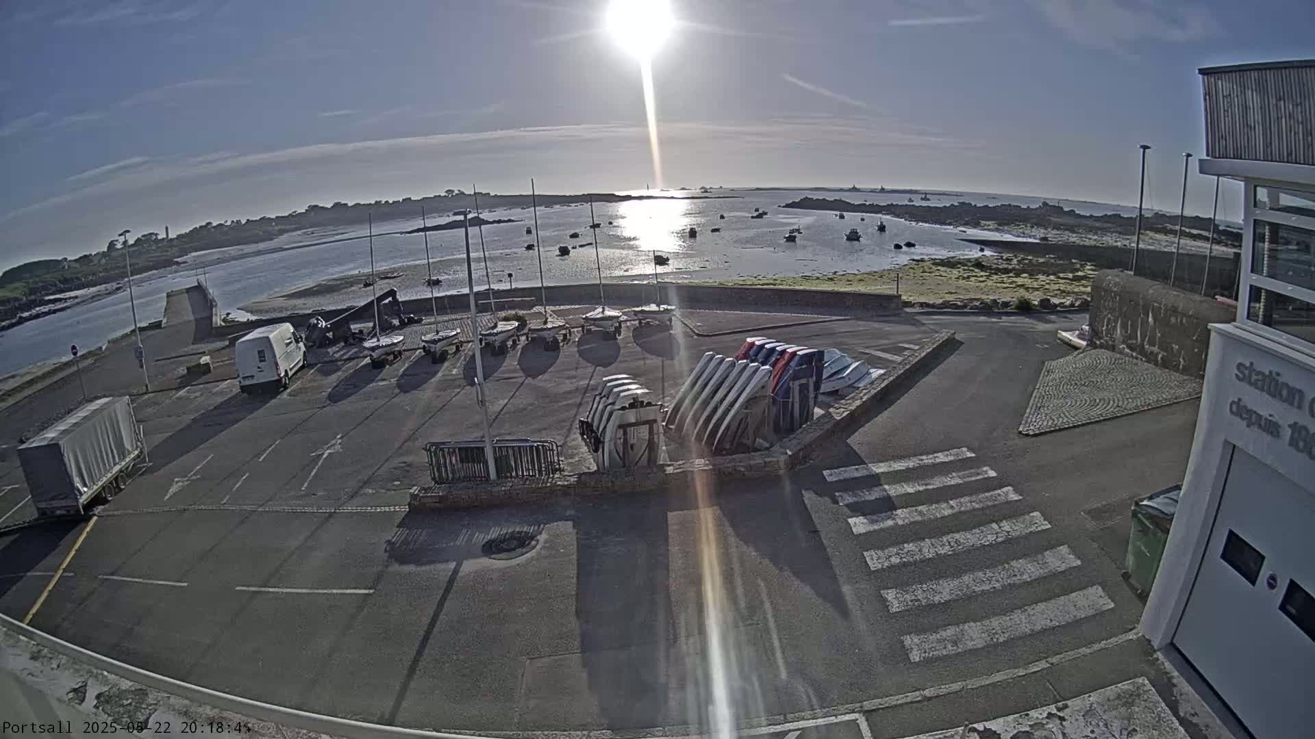 A sunny day at a harbor shows sailboats docked, several more stored upright on the ground, and a few vehicles parked nearby.