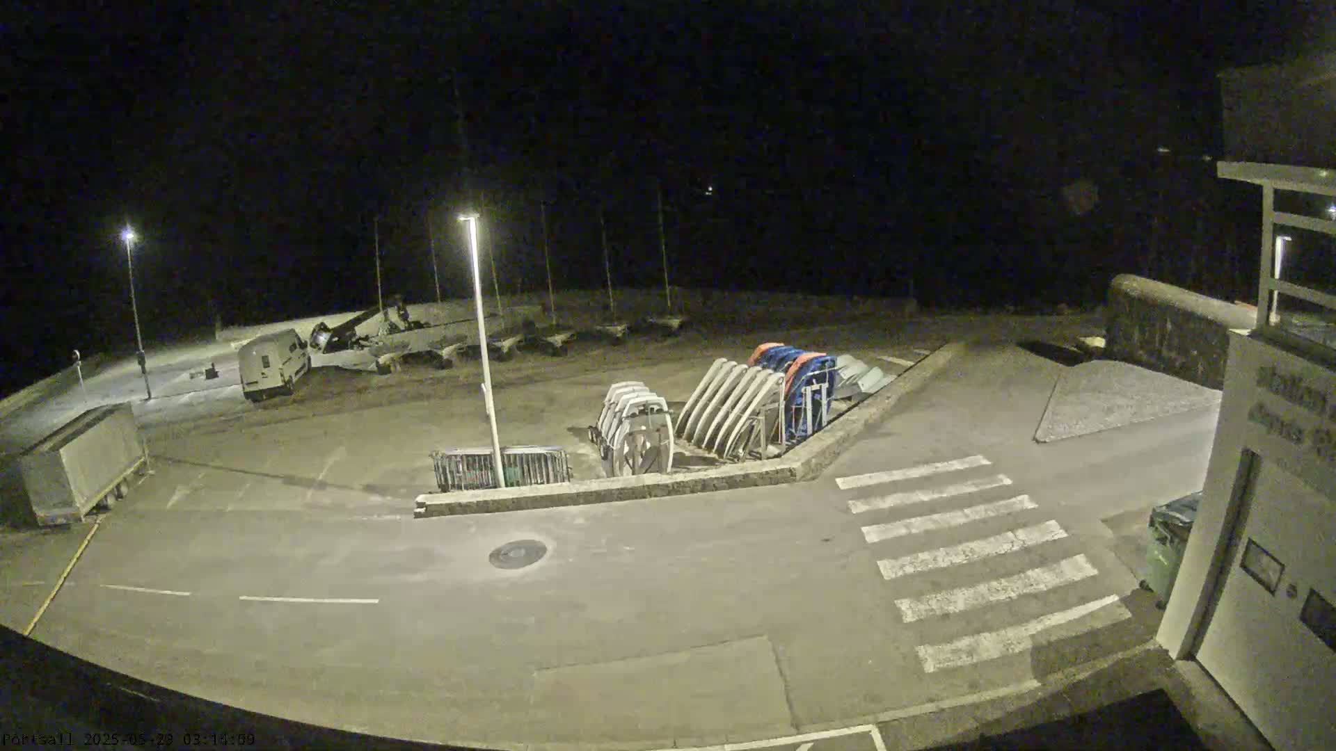 A nighttime, dimly lit parking lot contains several vehicles, boats on trailers, and a building, all under a dark sky.