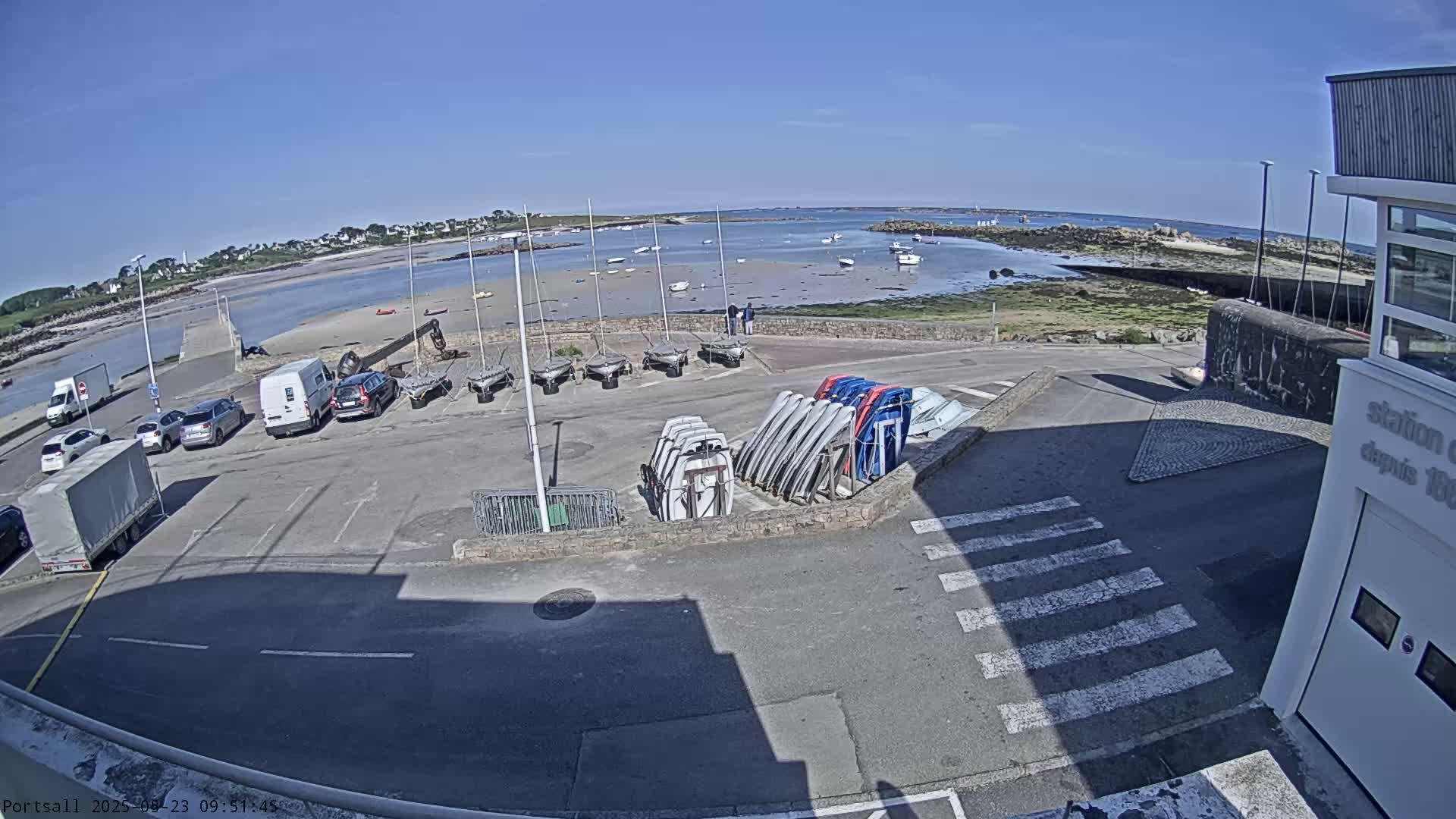 A sunny day at a harbor shows a parking lot with several cars and vans, along with numerous sailboats stored on racks, overlooking a calm body of water with boats anchored in the distance.