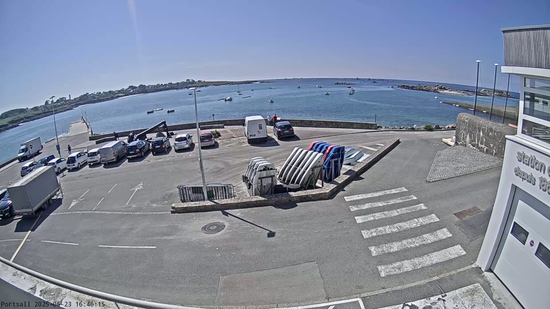 A parking lot with several cars and a few vans is next to a calm body of water with numerous boats under a clear, sunny sky.