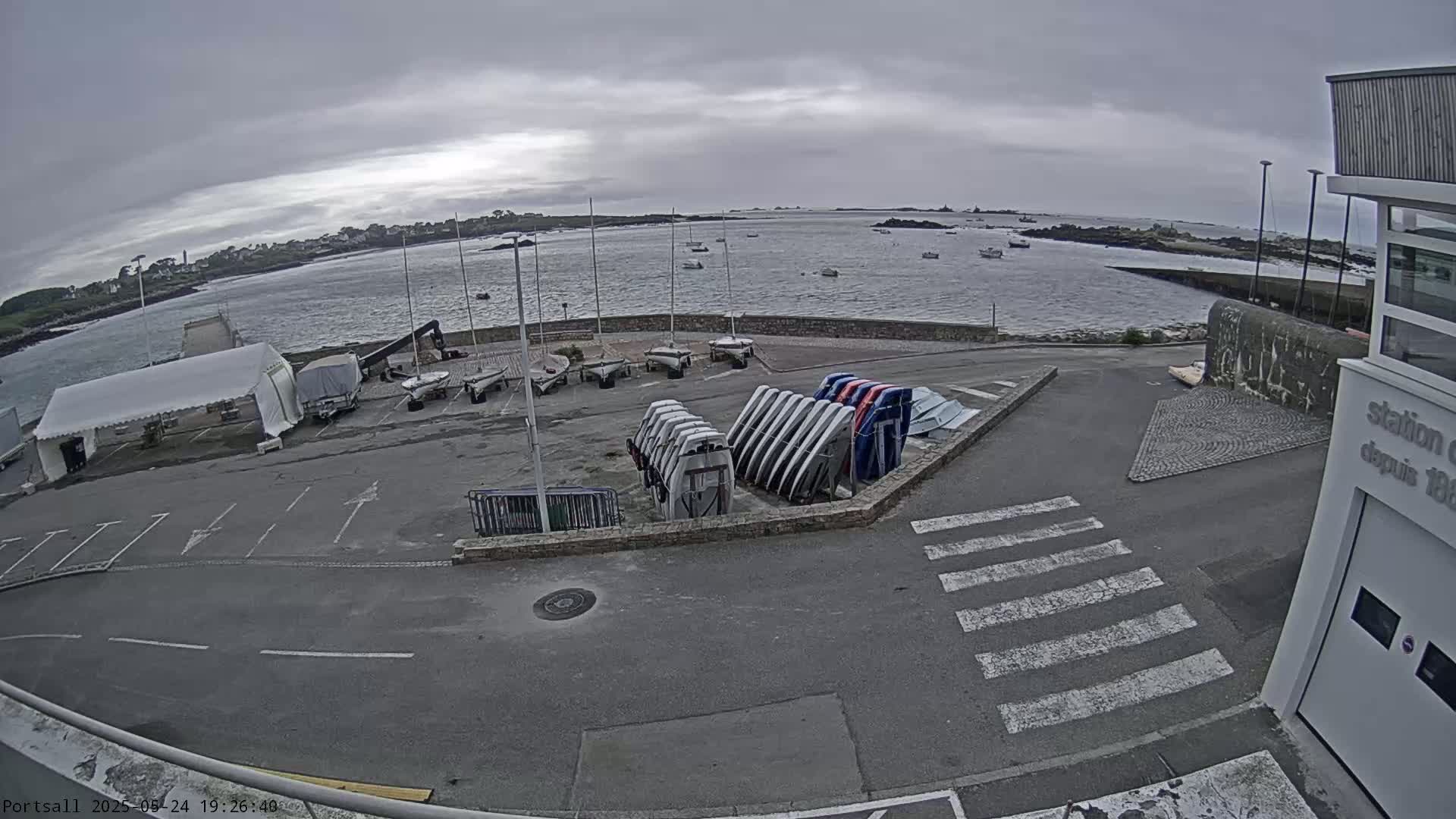 A parking lot with several sailboats and trailers is situated near a body of water under an overcast sky.