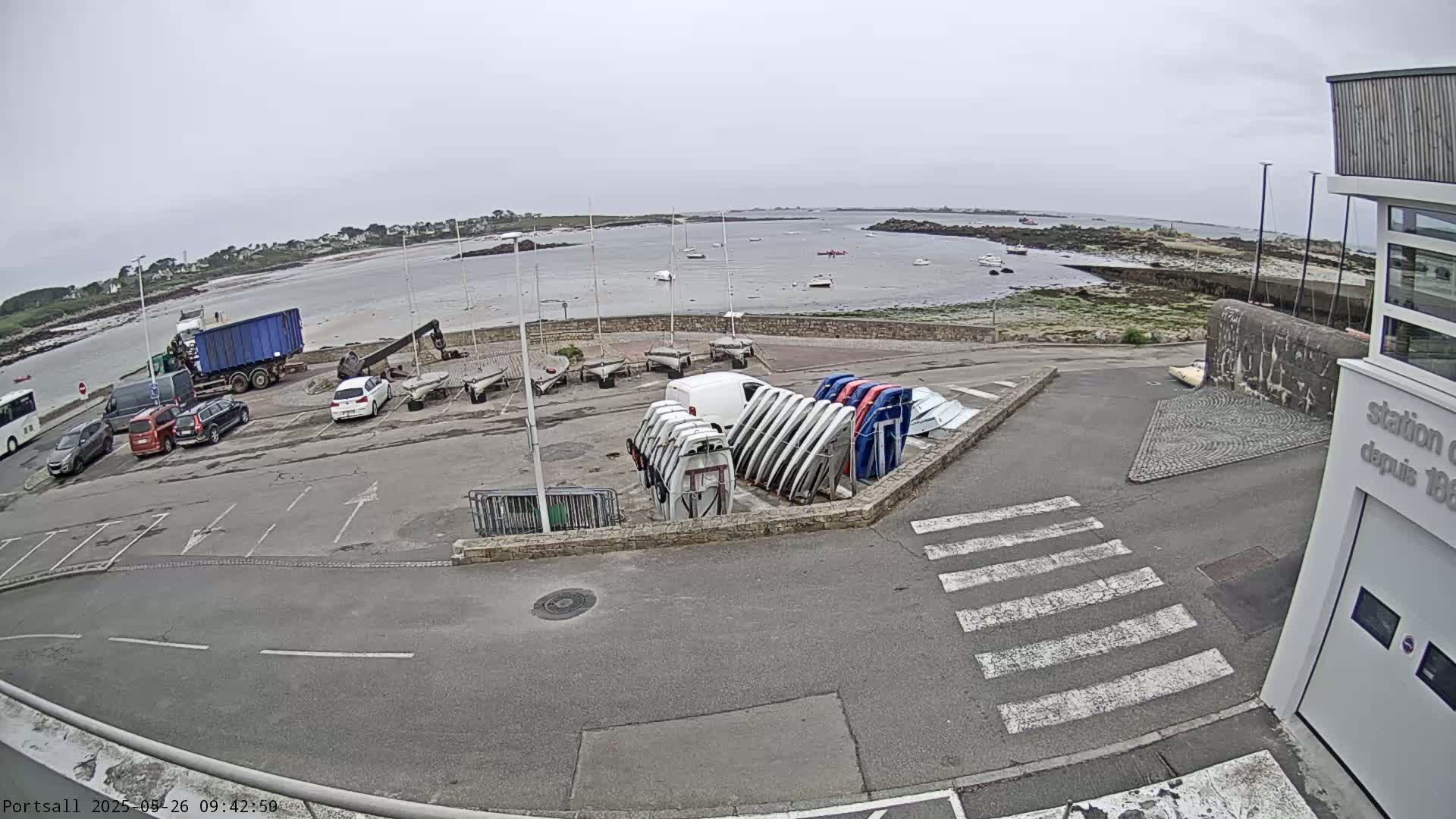 A parking lot with several cars and boats, some of which are stored upright, overlooks a calm body of water under an overcast sky.