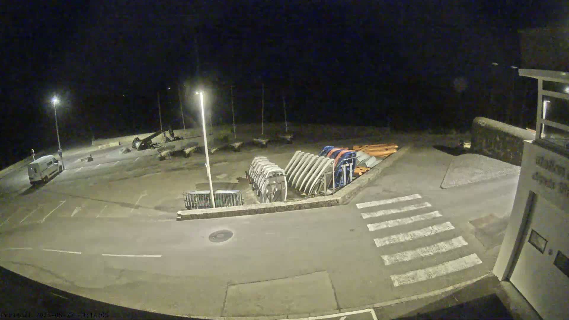 A nighttime, outdoor view of a parking lot with several boats on trailers, a van, and a pedestrian crossing.