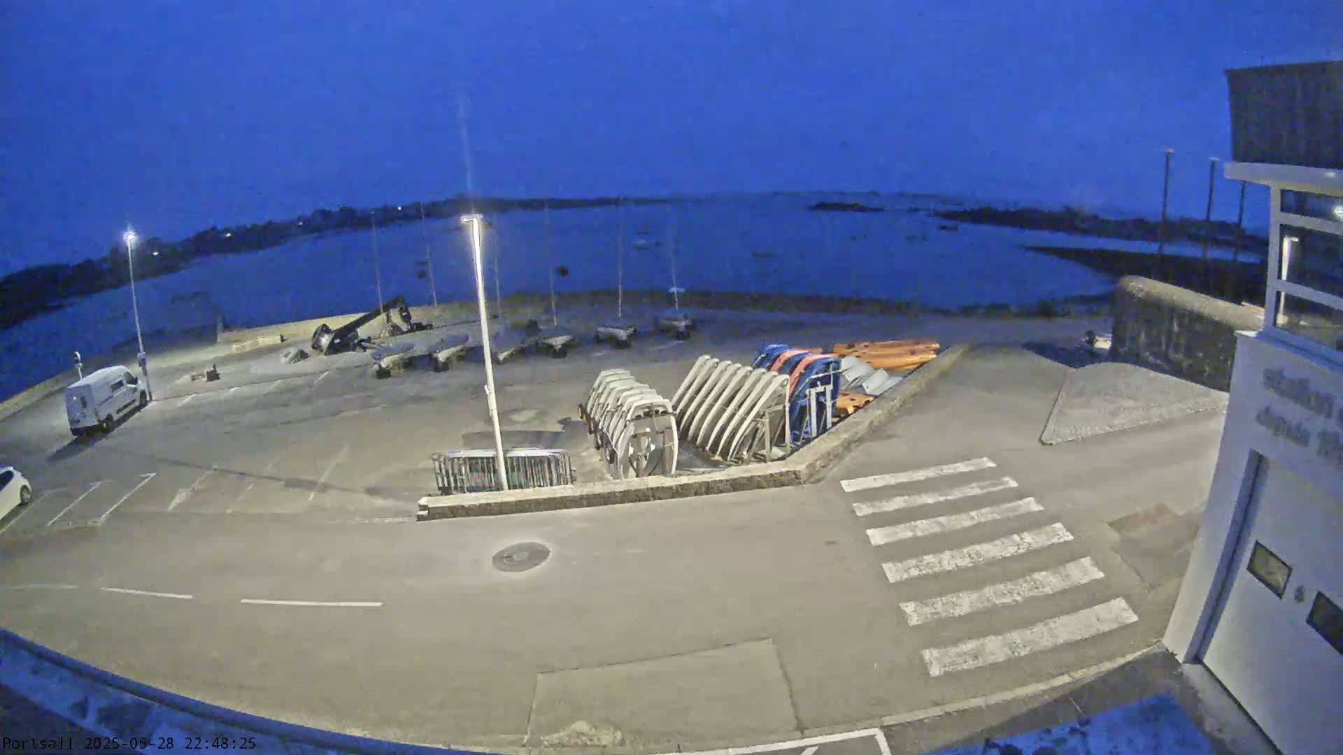 A nighttime view of a harbor with a parking lot containing several boats on trailers, a van, and a small building.
