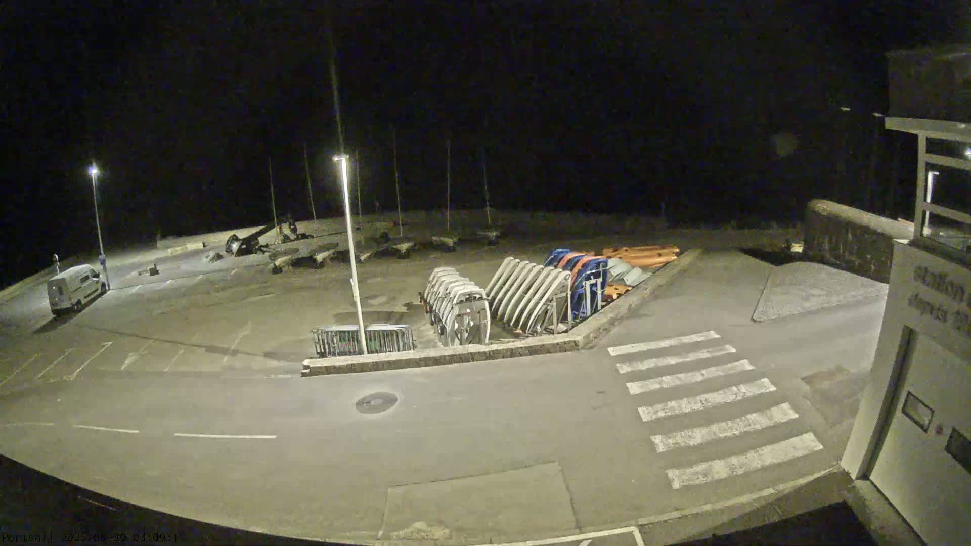 A nighttime, overhead view of a mostly empty parking lot with several boats on trailers, a van, and a crosswalk.
