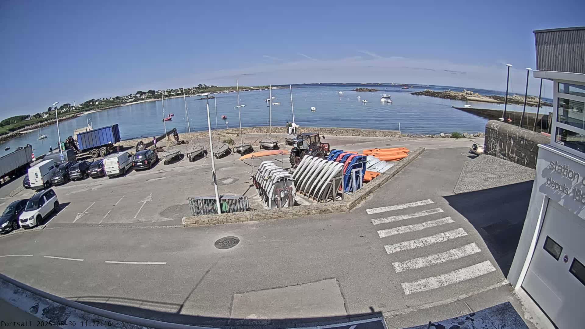A sunny day at a harbor shows a parking lot with several cars and trucks, a collection of sailboats and kayaks, and a calm body of water with boats in the distance.