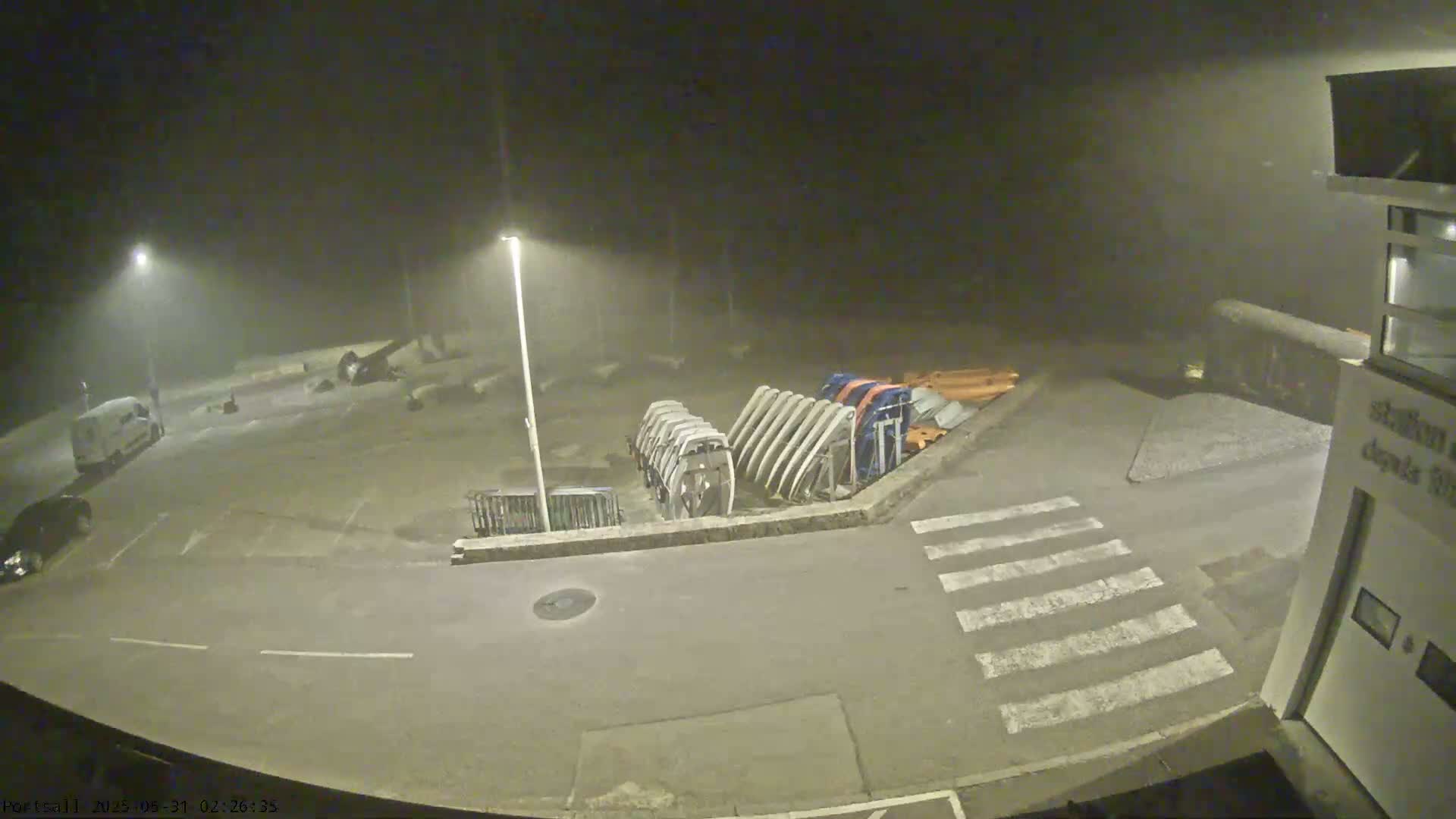 A foggy night scene shows a mostly empty parking lot with a few vehicles, a group of boat hulls, and a crosswalk.
