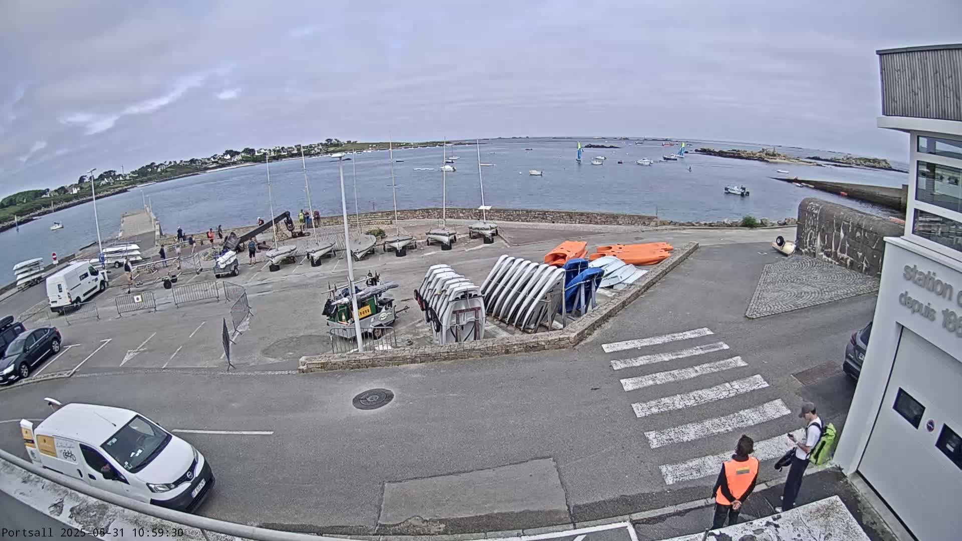 A harbor with numerous small sailboats and other watercraft, along with several parked vehicles and two people standing near a building, under overcast skies.