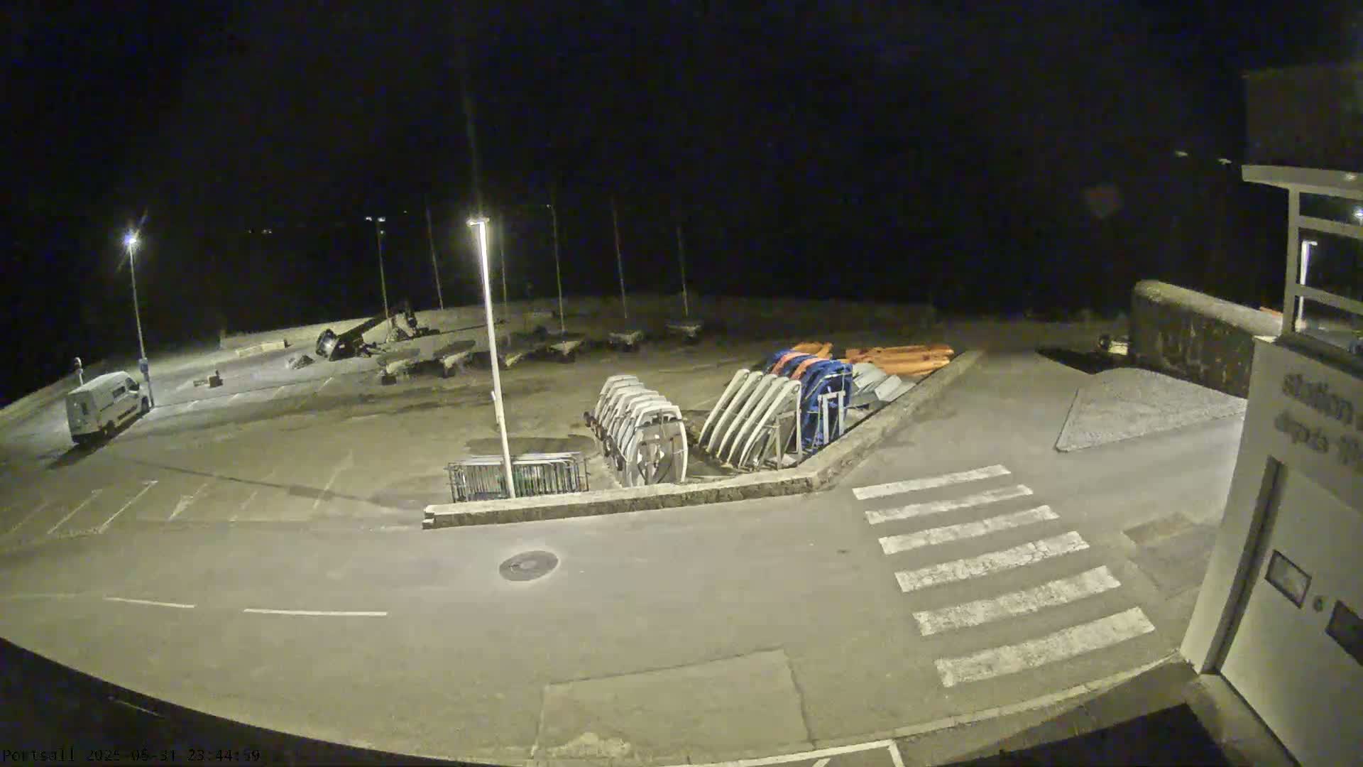 At night, an empty parking lot next to a body of water contains several boats and trailers under bright lights.