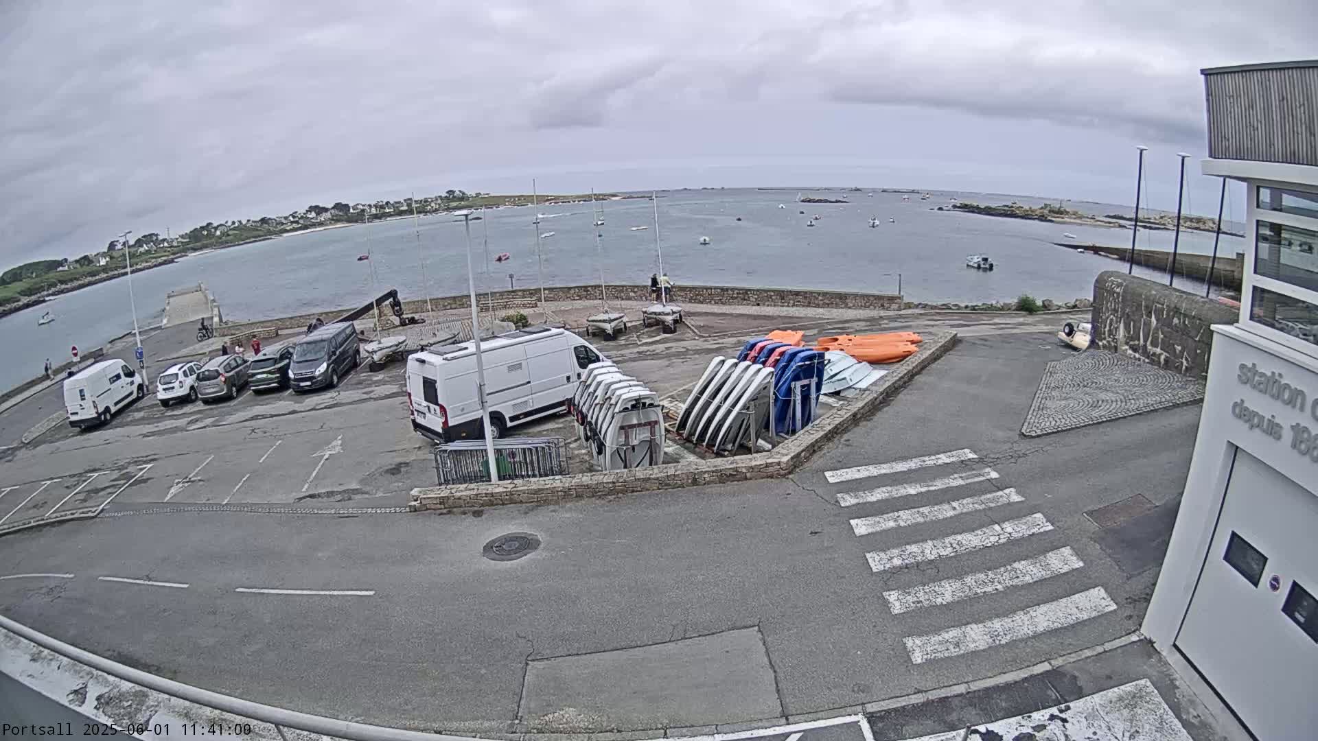 A parking lot with several cars and a van, next to a building and a harbor with many boats on a cloudy day.