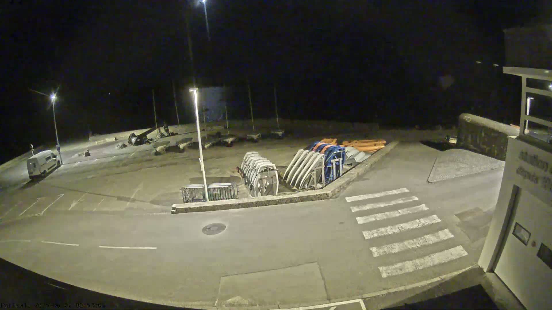 A nighttime, outdoor view of a mostly empty parking lot with several boats and boat trailers stored near a building.
