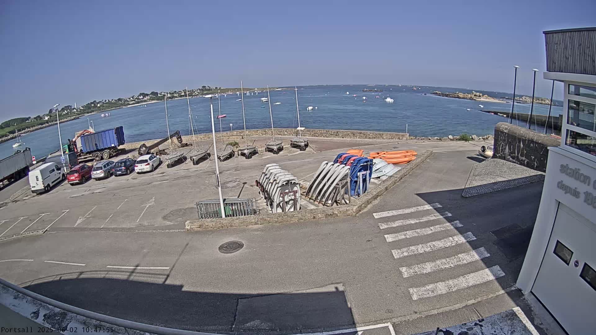 A sunny day at a harbor shows a parking lot with several cars and a large number of sailboats and other small boats stored on the ground, with a body of water in the background.