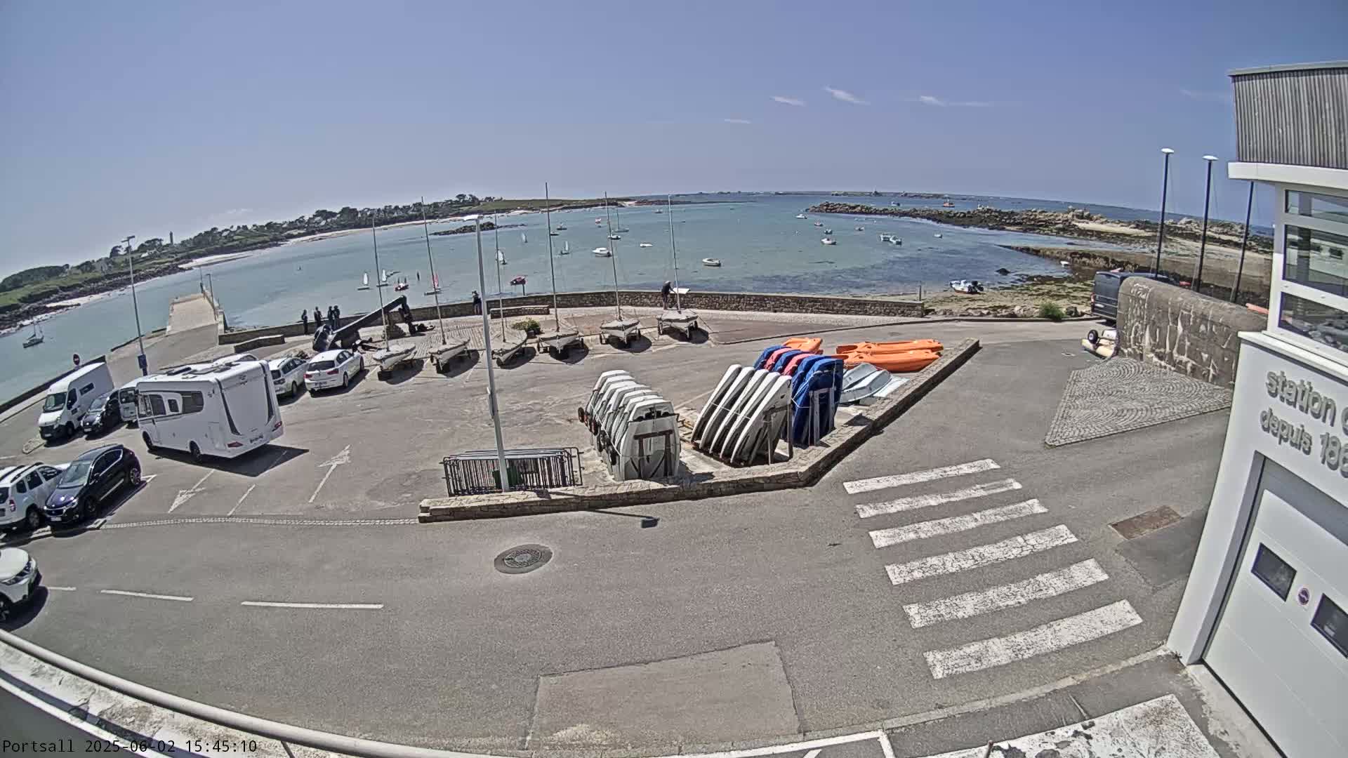 A sunny day at a harbor shows sailboats on the water, parked cars and camper vans, and numerous small boats and kayaks stored on land.