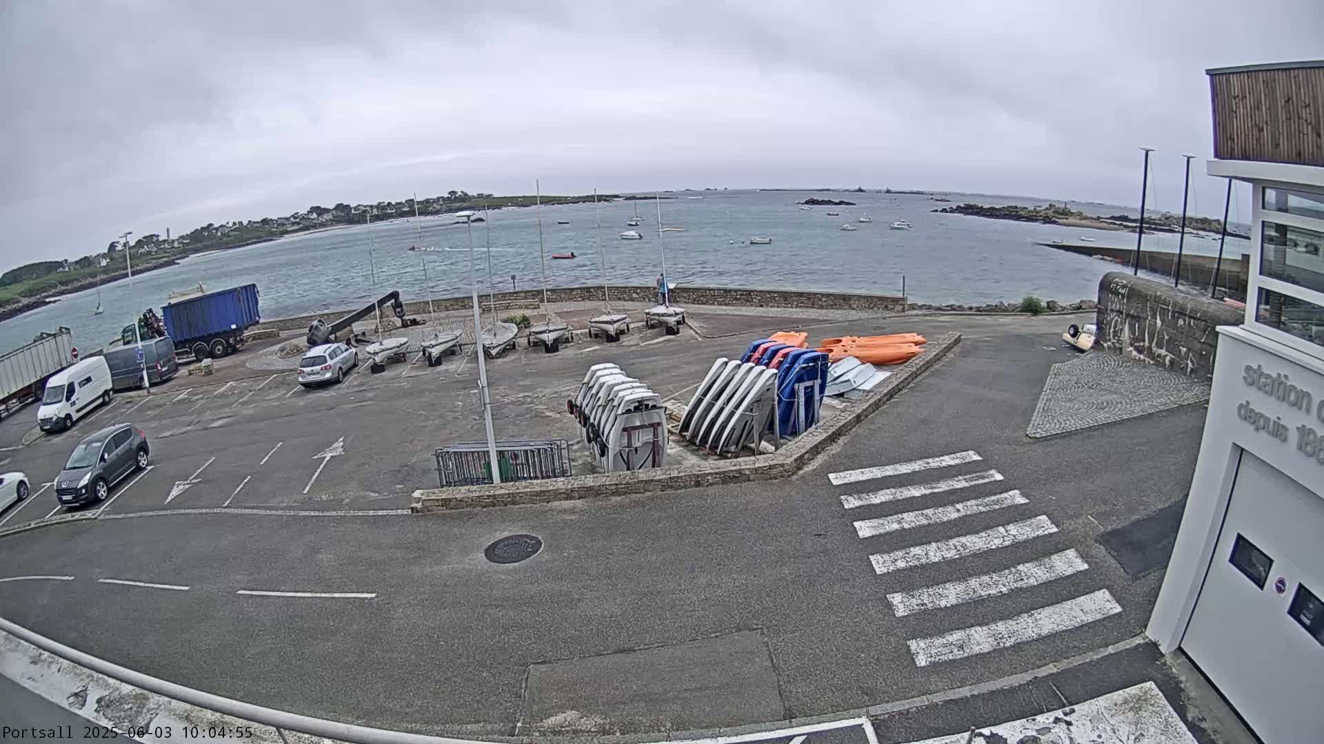 A parking lot with several cars and trucks is adjacent to a calm body of water under an overcast sky, with numerous sailboats and other small watercraft docked or stored nearby.